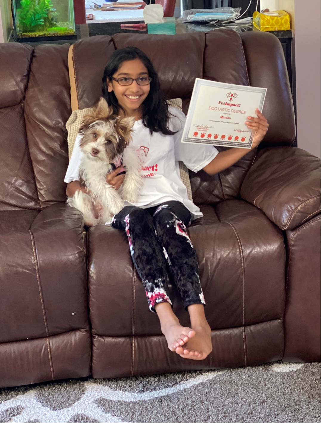 Girl with small dog on brown recliner, holding a certificate, smiling.