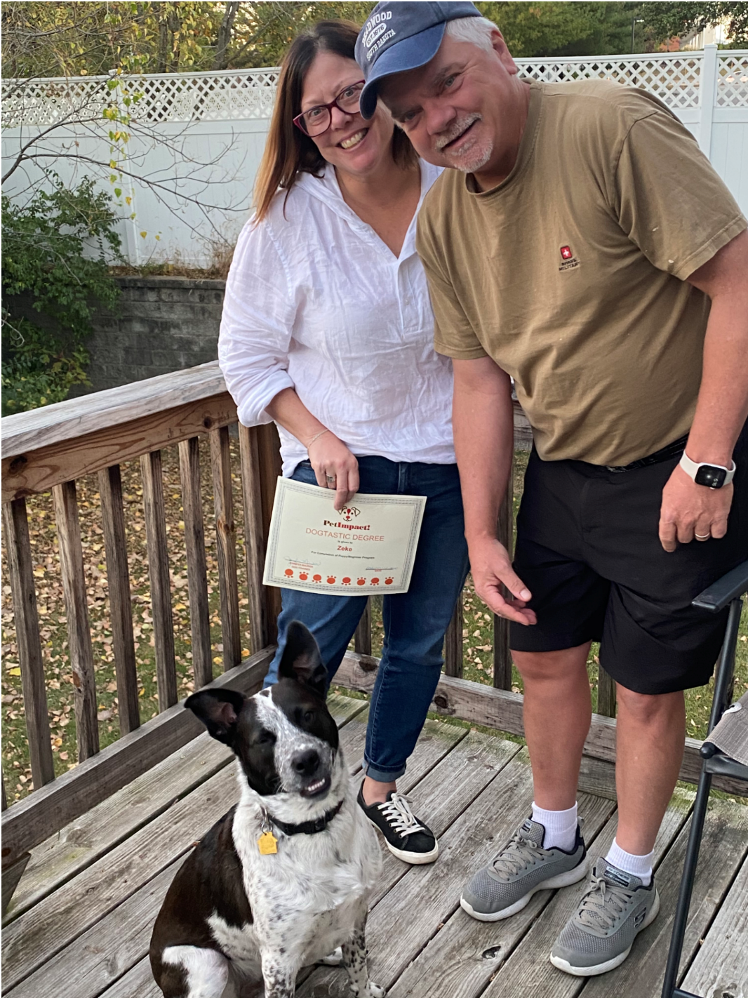 A couple and a dog on a deck. The woman holds a certificate. The dog sits and smiles.