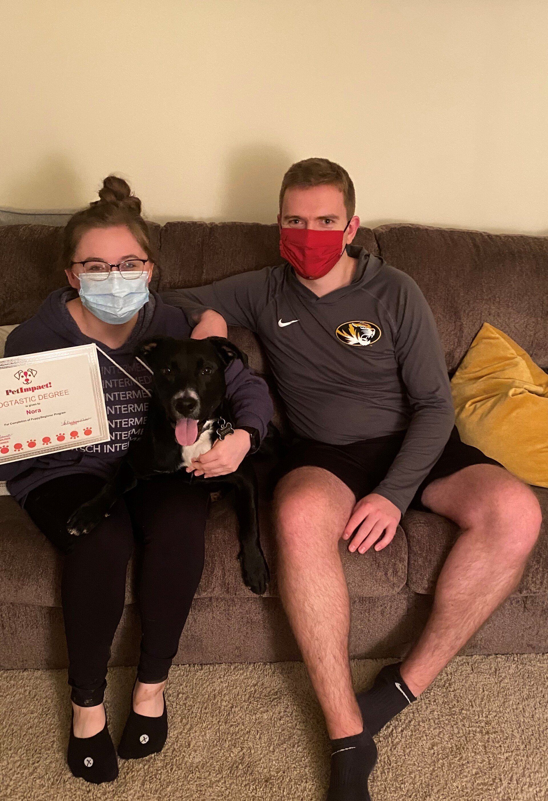 Two people and a dog seated on a couch; all wearing masks. One person holds a sign.
