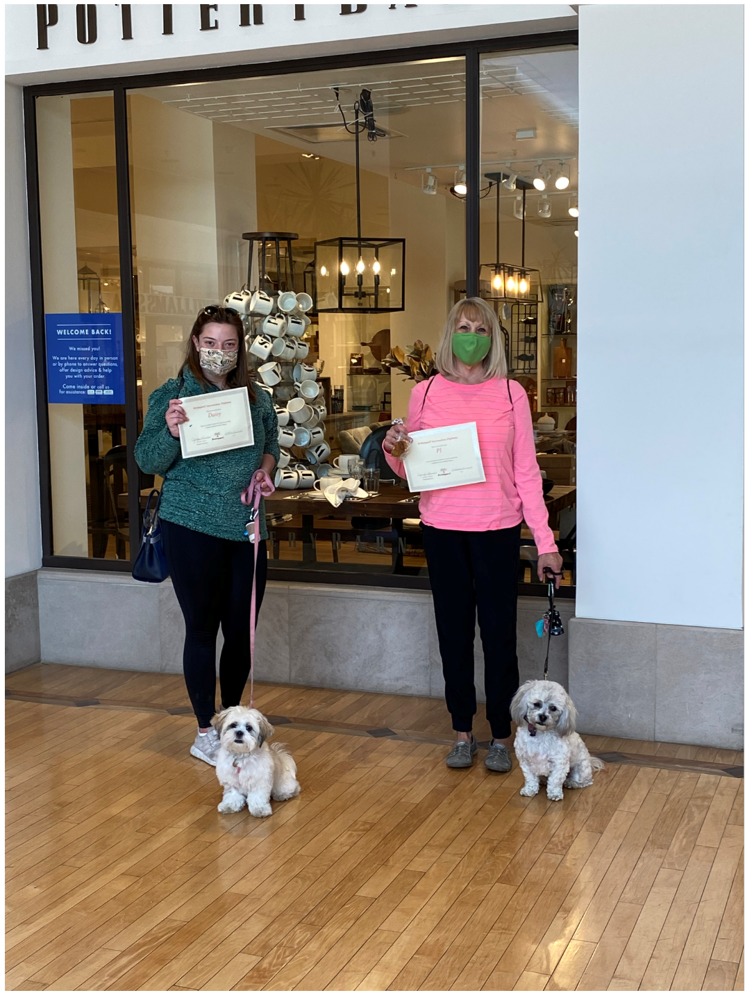 Two women with small white dogs in a store doorway, holding papers. One woman wears a green mask, pink sweater.