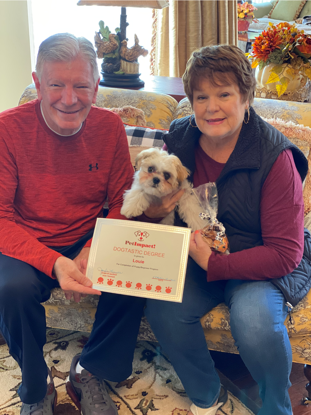 Couple holding a puppy and a certificate, seated indoors. Man in red shirt, woman in vest.