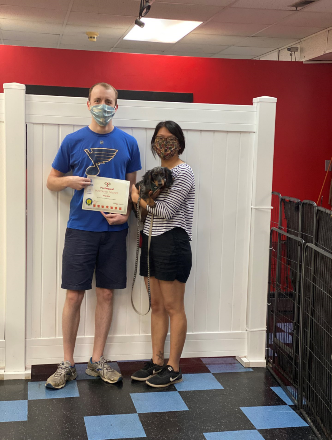 Man holding certificate and woman holding small dog, both masked, in front of a white fence.