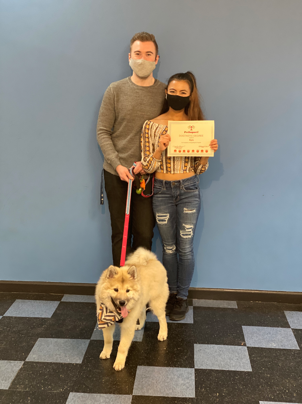 Man and woman with dog, holding a certificate in front of a blue wall. The dog wears a bandana and leash.