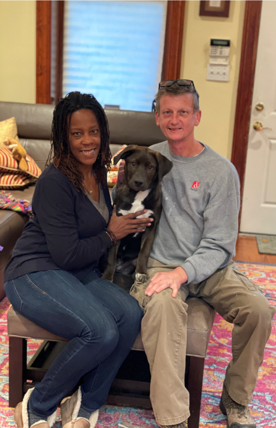 Couple seated, holding a black and white dog. They're indoors. The woman is wearing a dark blue sweater, jeans, and sneakers. The man has a gray shirt and tan pants.