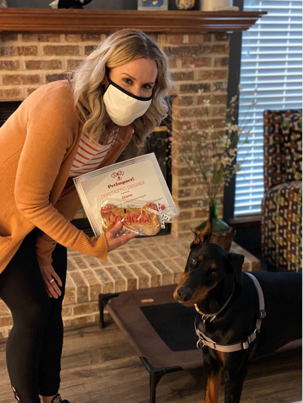 Woman in mask holding dog treats, standing with a Doberman. Brick fireplace in the background.