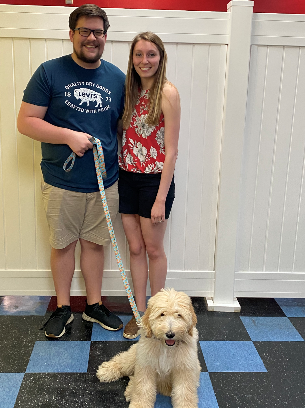 Couple with golden doodle, standing indoors with a white wall. The dog sits facing forward, smiling.