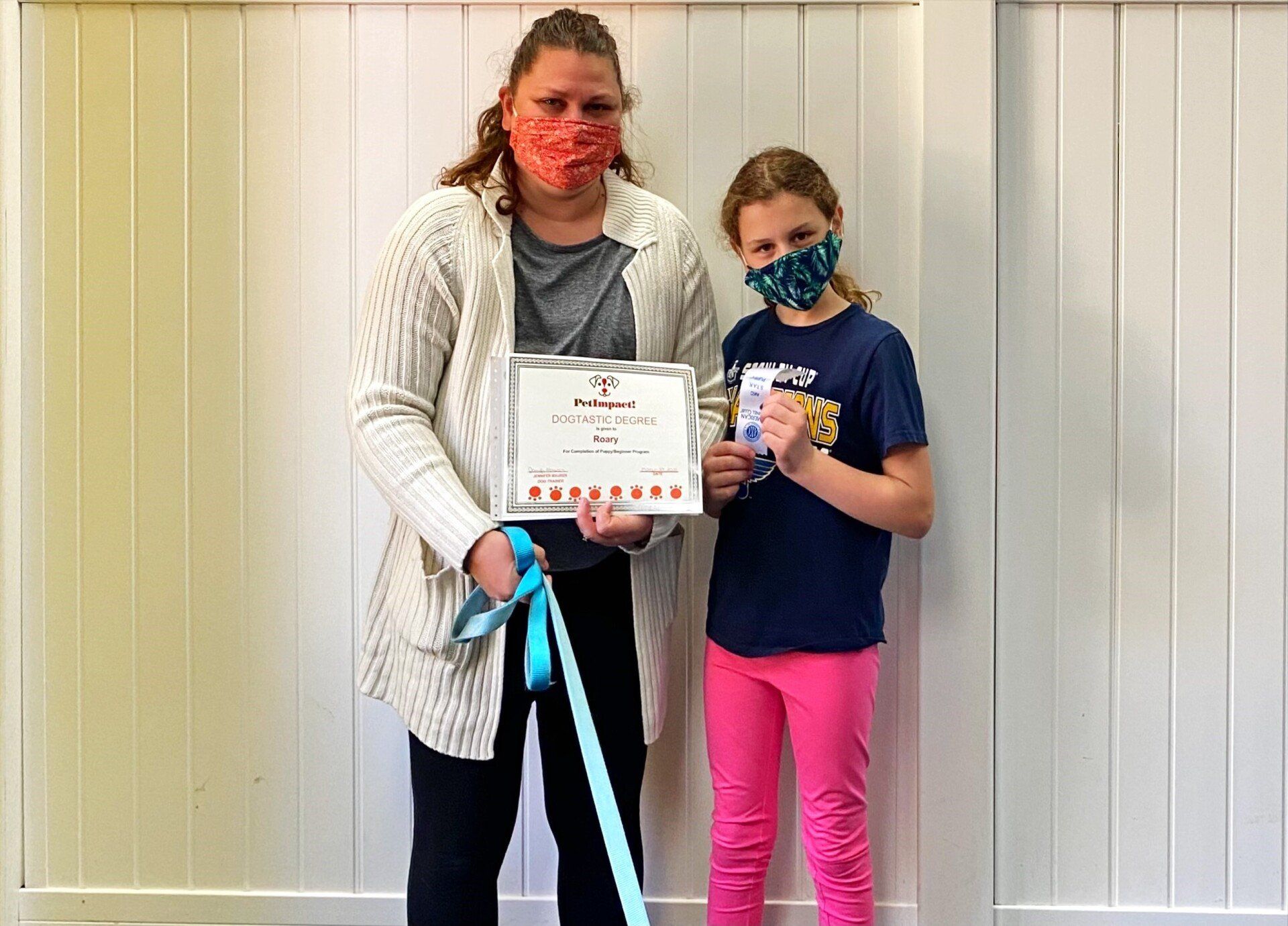 Woman and girl holding certificate, both wearing masks, in front of a white wall.