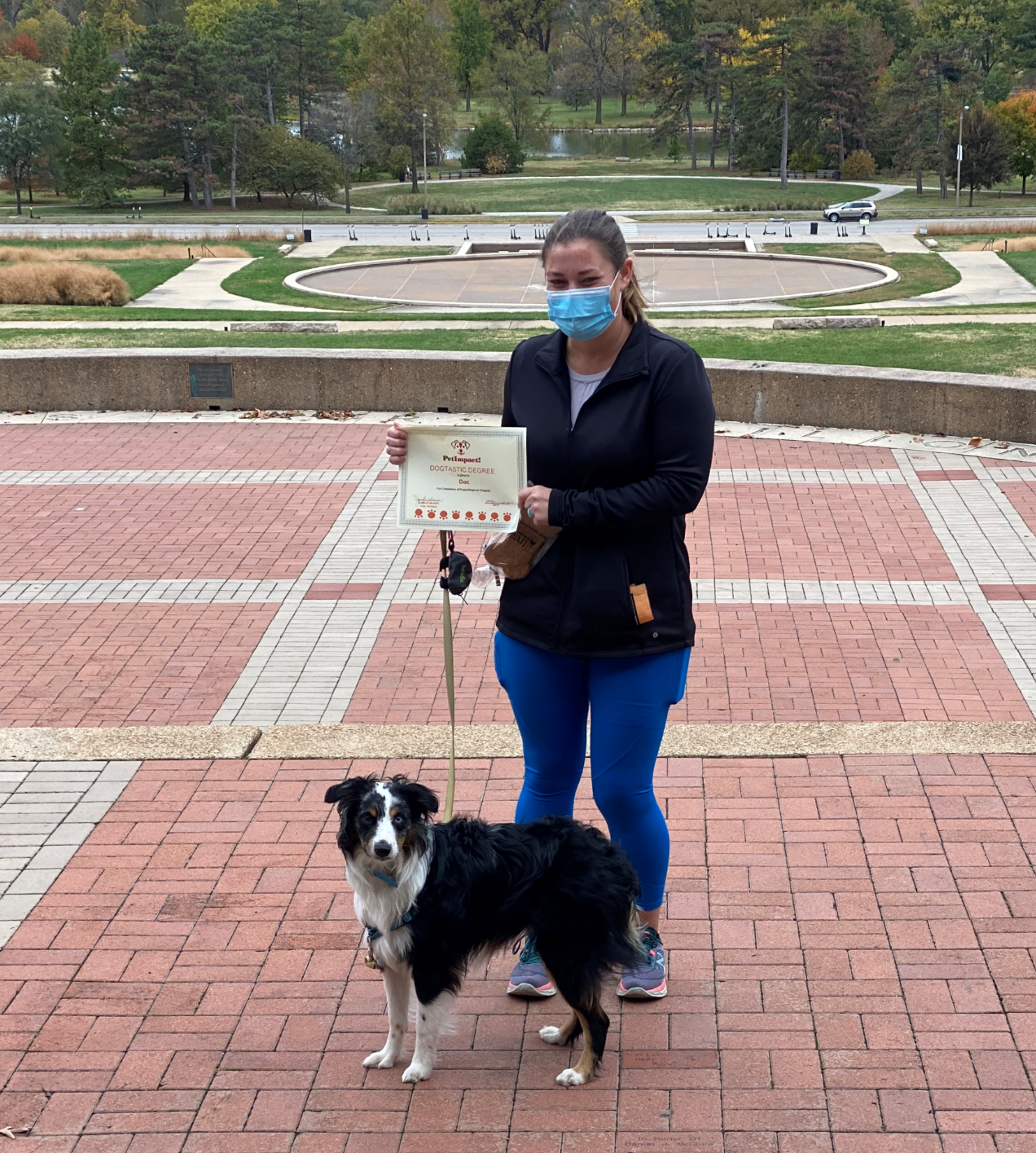 Woman in a mask holds a certificate next to a black and white dog in an outdoor setting.
