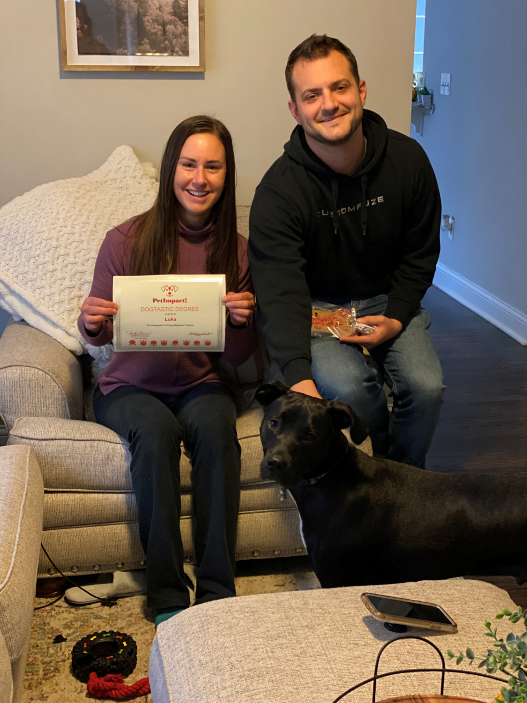 Woman holding a certificate, man with dog and snacks, sitting on a sofa in a living room.