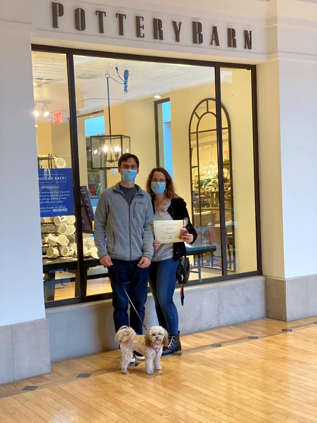 Couple with two small dogs in front of Pottery Barn store. They wear masks and hold a document.