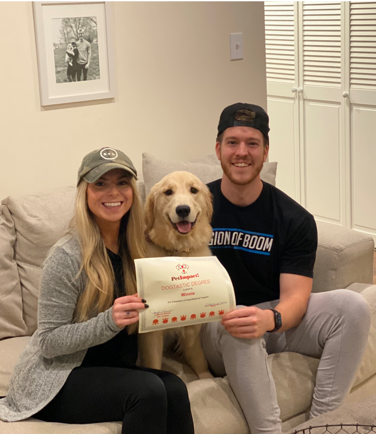 Couple and golden retriever pose indoors, holding certificate. Dog smiles, people are happy.
