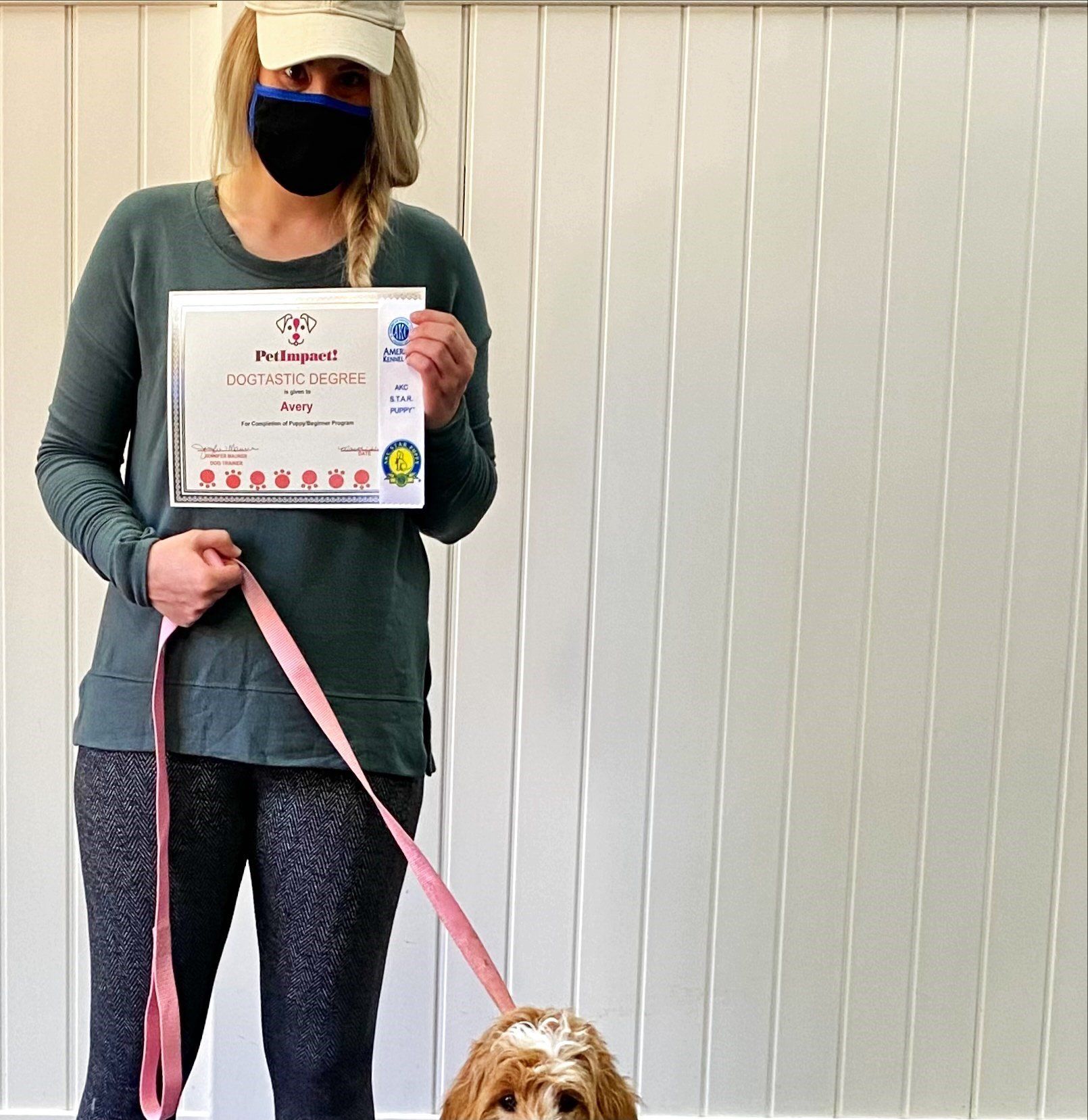 Woman in mask holding a certificate, standing with a dog on a leash in front of a white shuttered wall.