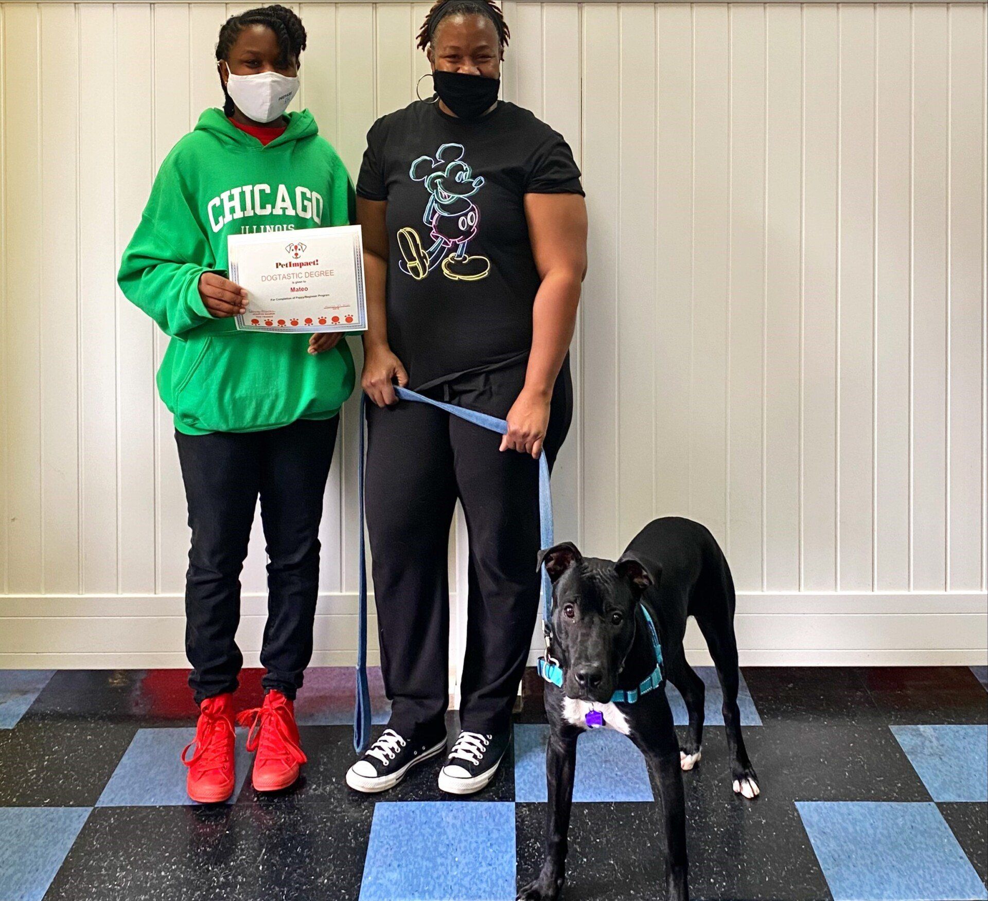 Two people with a dog, one holding a certificate. A person in green holds the certificate, another in black holds the leash. A black dog stands in front of them.