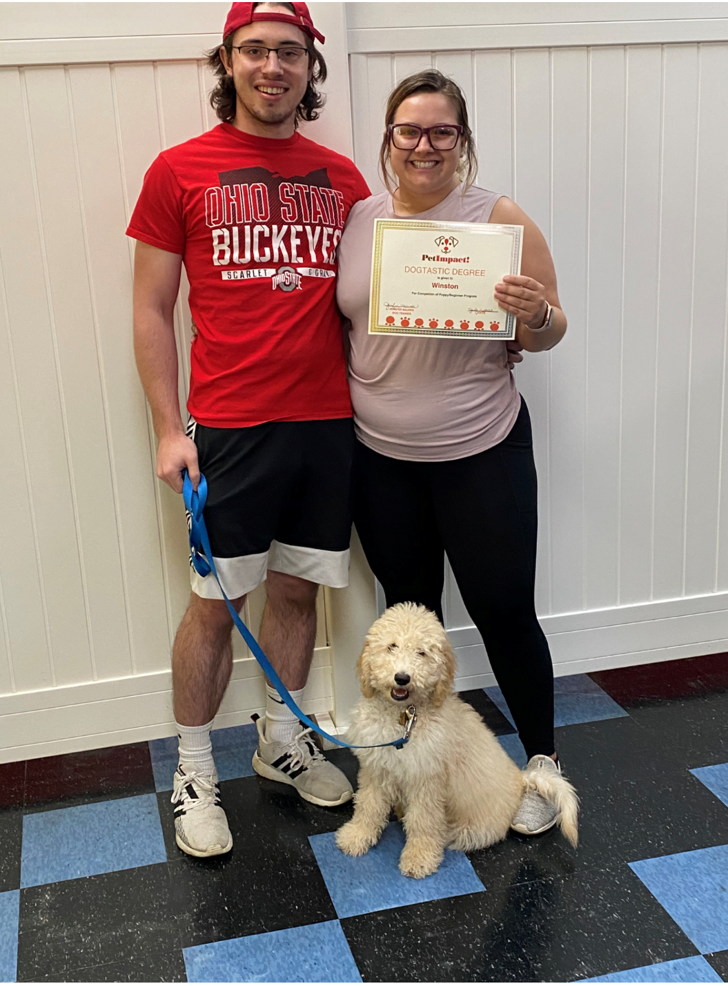 Man and woman with dog; woman holds a certificate. They pose against a white wall and blue tiled floor.