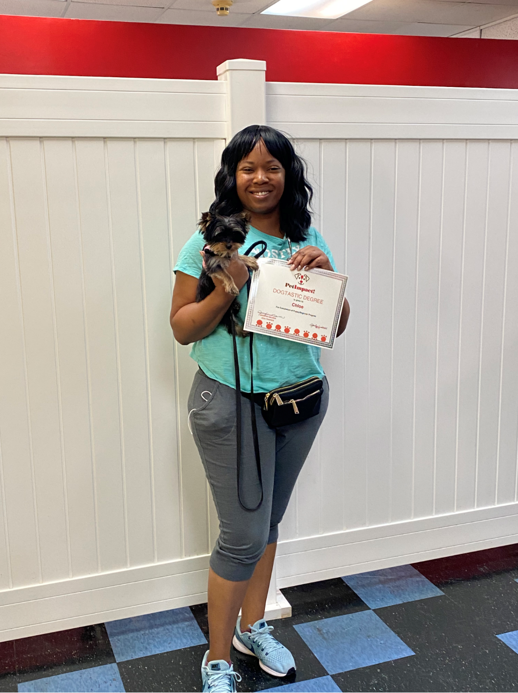 Woman holding small dog and certificate, standing in front of a white wall with red trim.