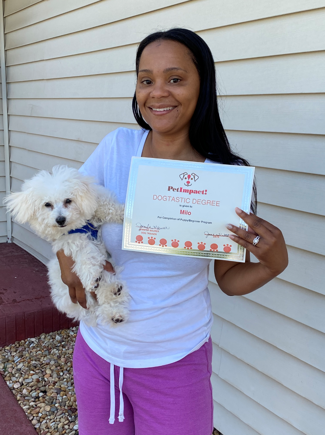 Woman holding a small white dog and a certificate outdoors, smiling.
