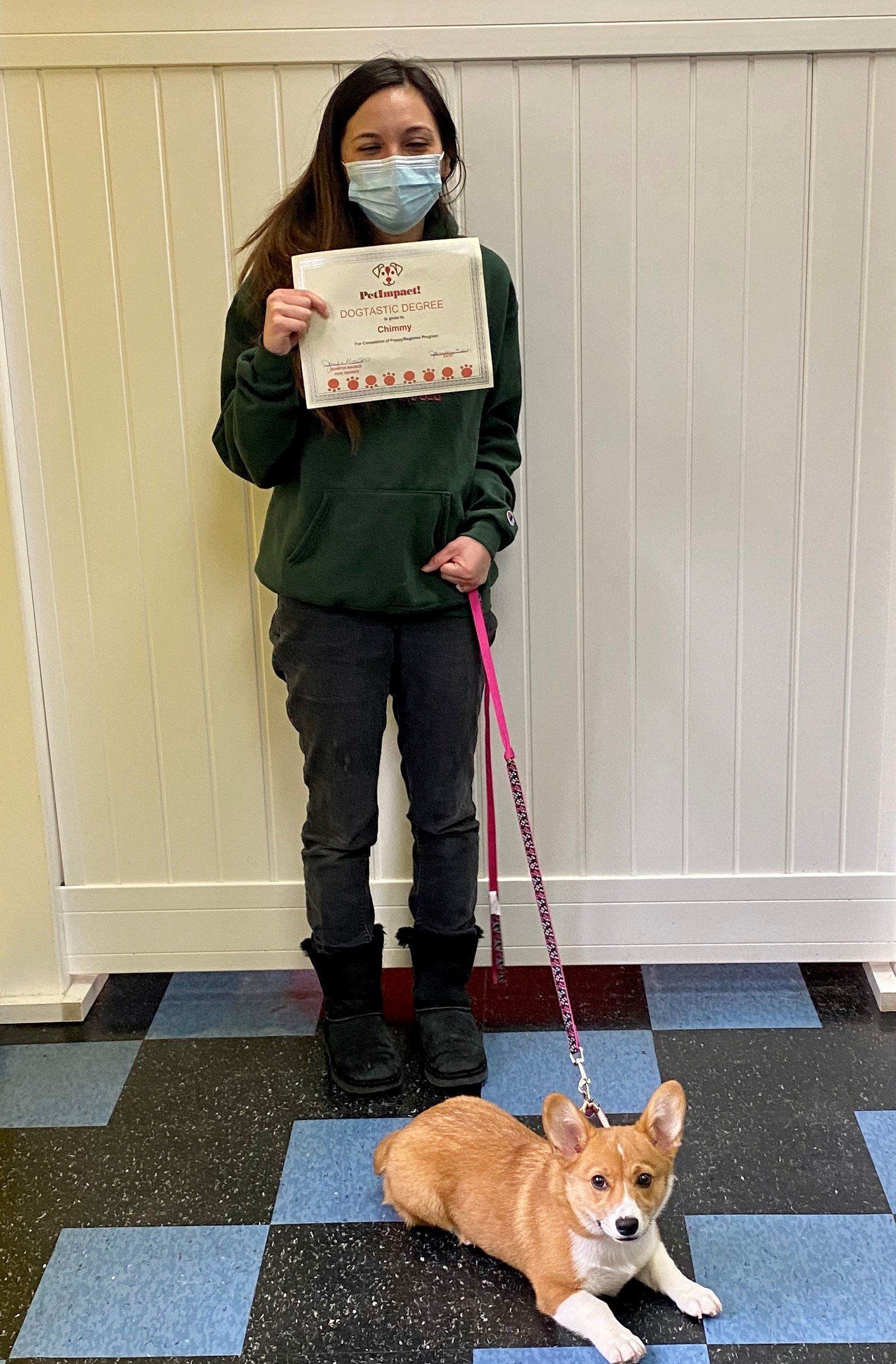 Woman in green hoodie holds sign, sitting corgi on leash.