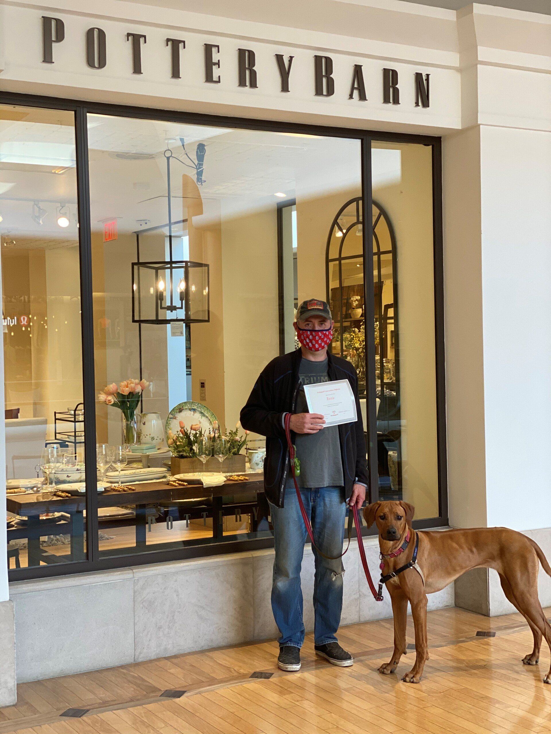 Man and dog in front of Pottery Barn store. Man wears a mask, holding a paper. Dog is on a leash.