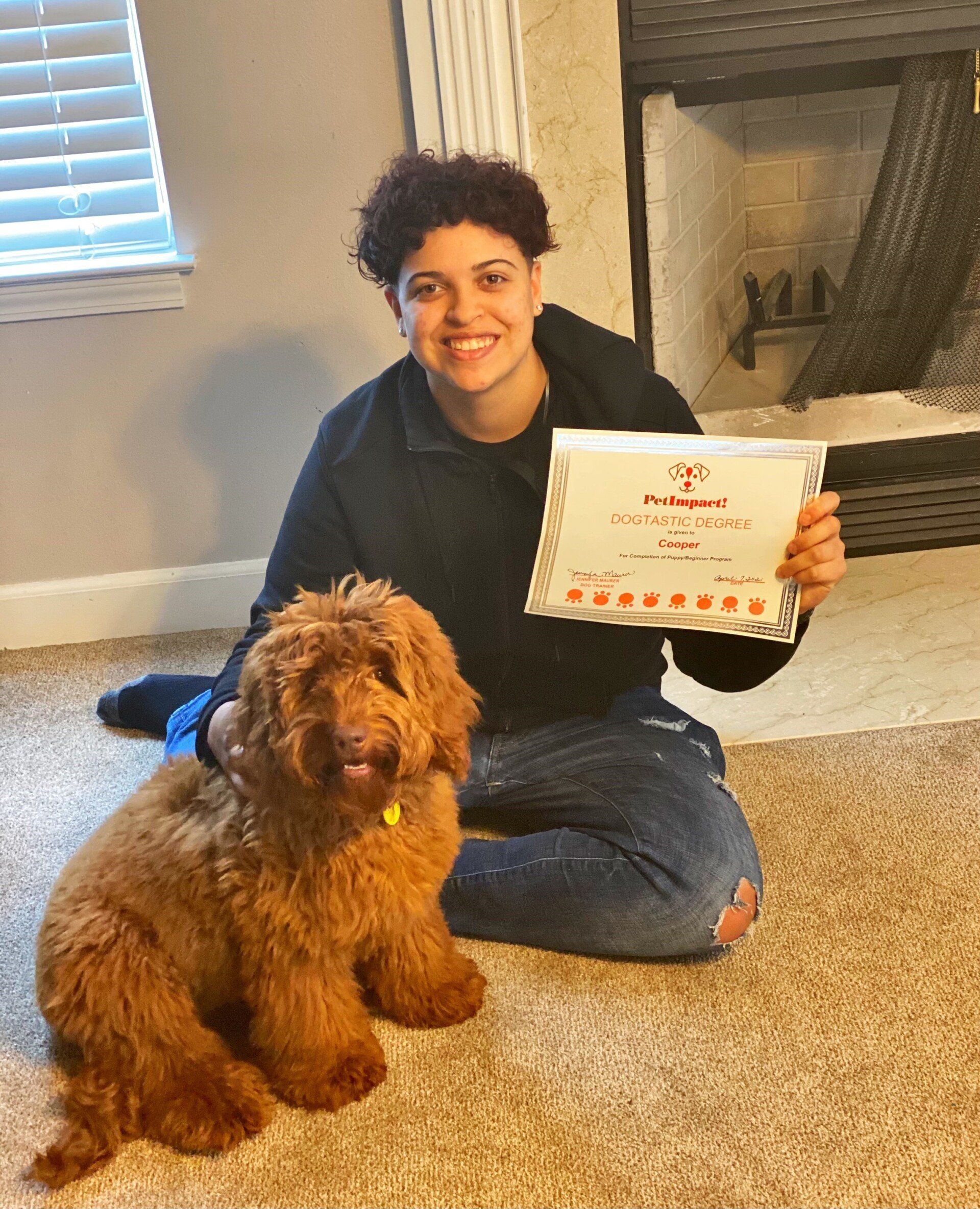 Person kneeling with a golden doodle dog, holding a certificate indoors near a fireplace.