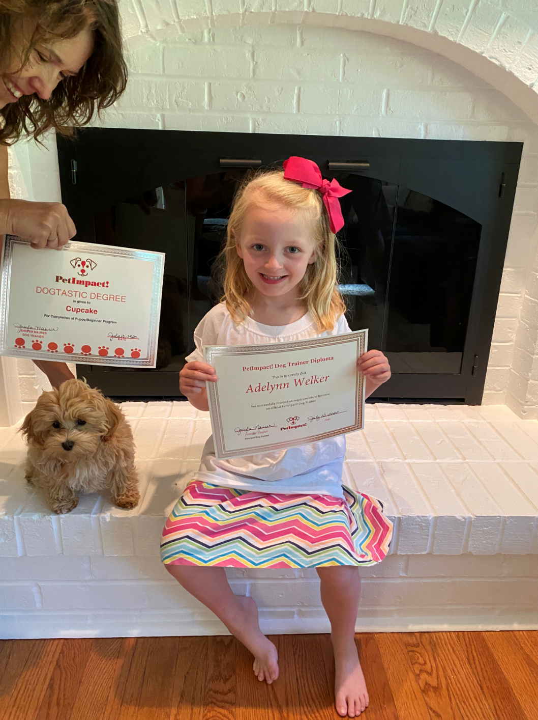 Girl sitting in front of a fireplace, holding a certificate. A woman and a toy dog are nearby.