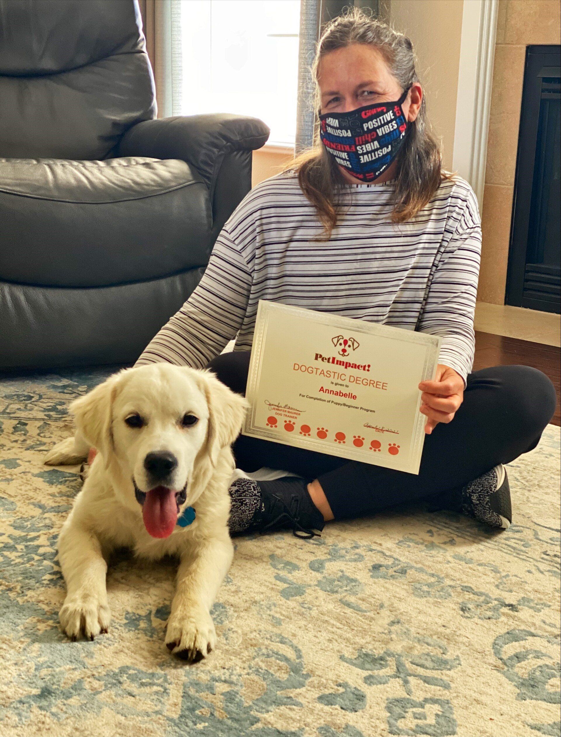 Woman with mask and golden retriever puppy, holding a certificate, sitting on the floor.