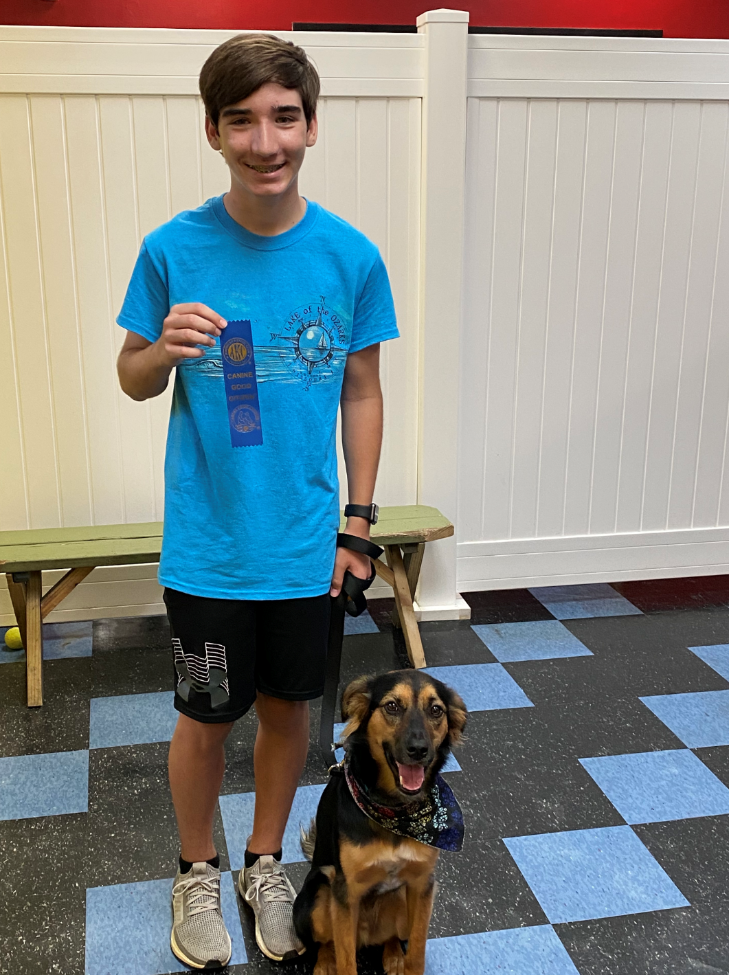 Teen with blue ribbon, smiling, standing with dog in indoor setting.