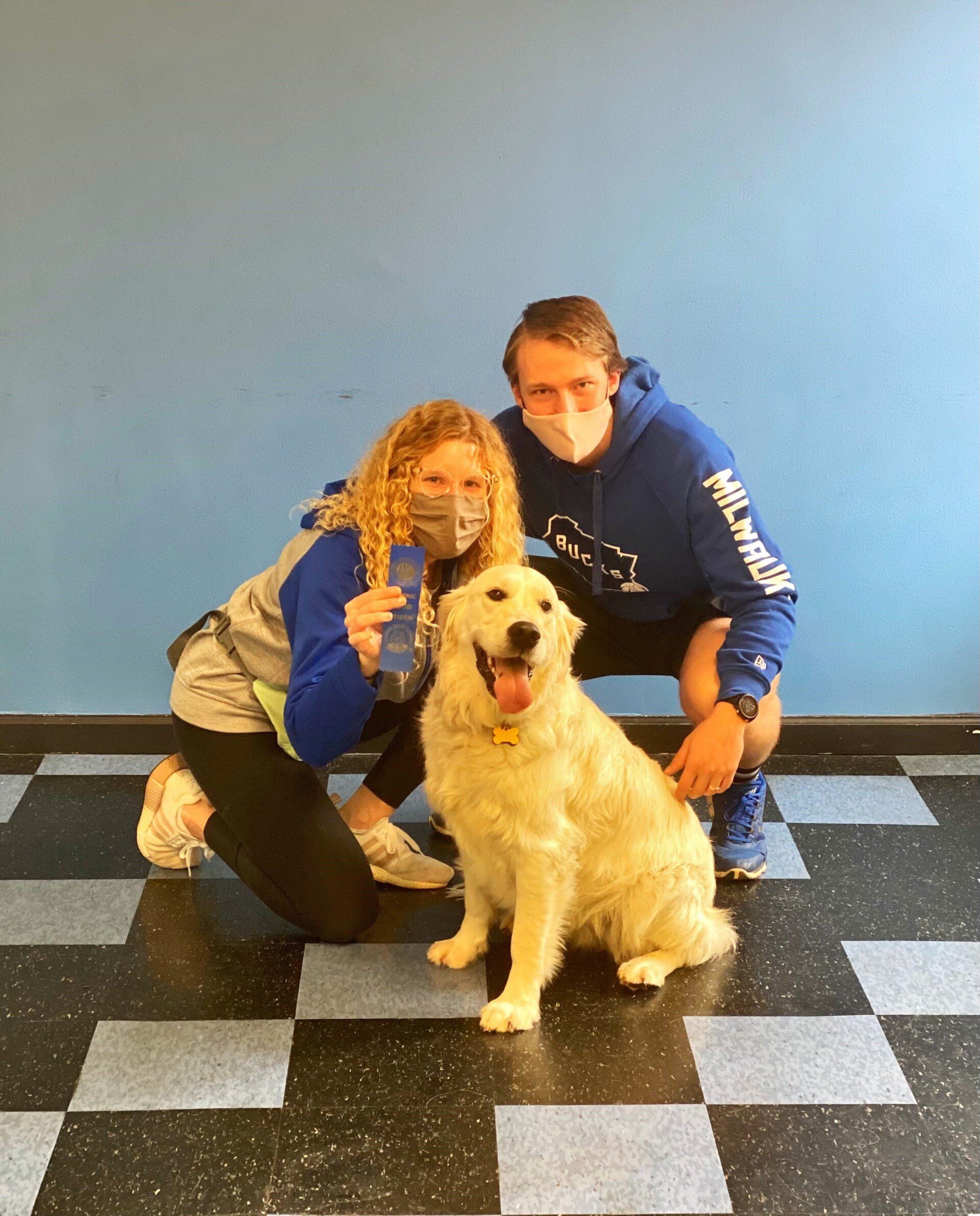 A golden retriever sits between two people in masks. They are on a checkered floor in front of a blue wall.