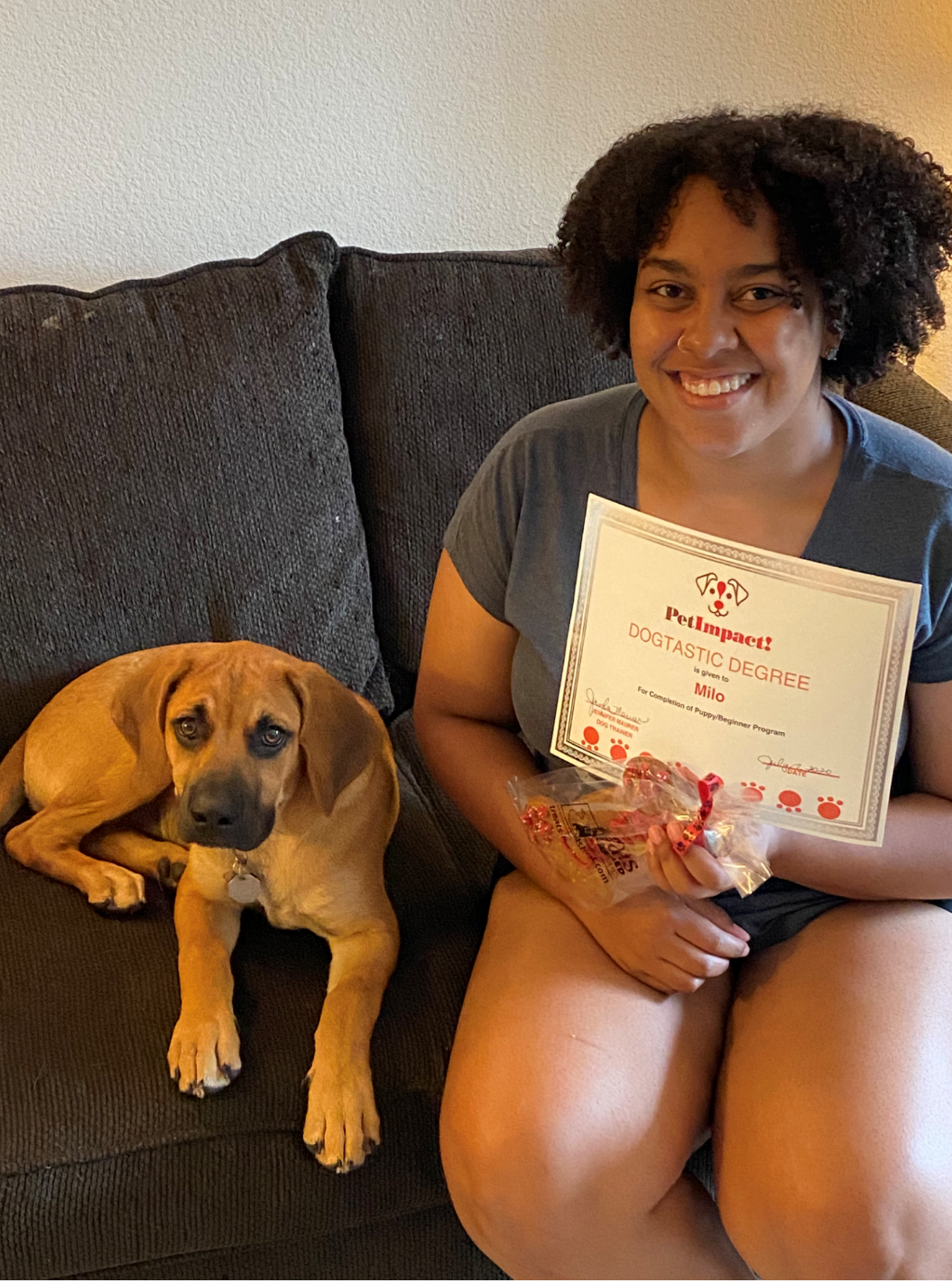 Woman on a couch, smiling, holding a certificate next to a brown dog; indoors.