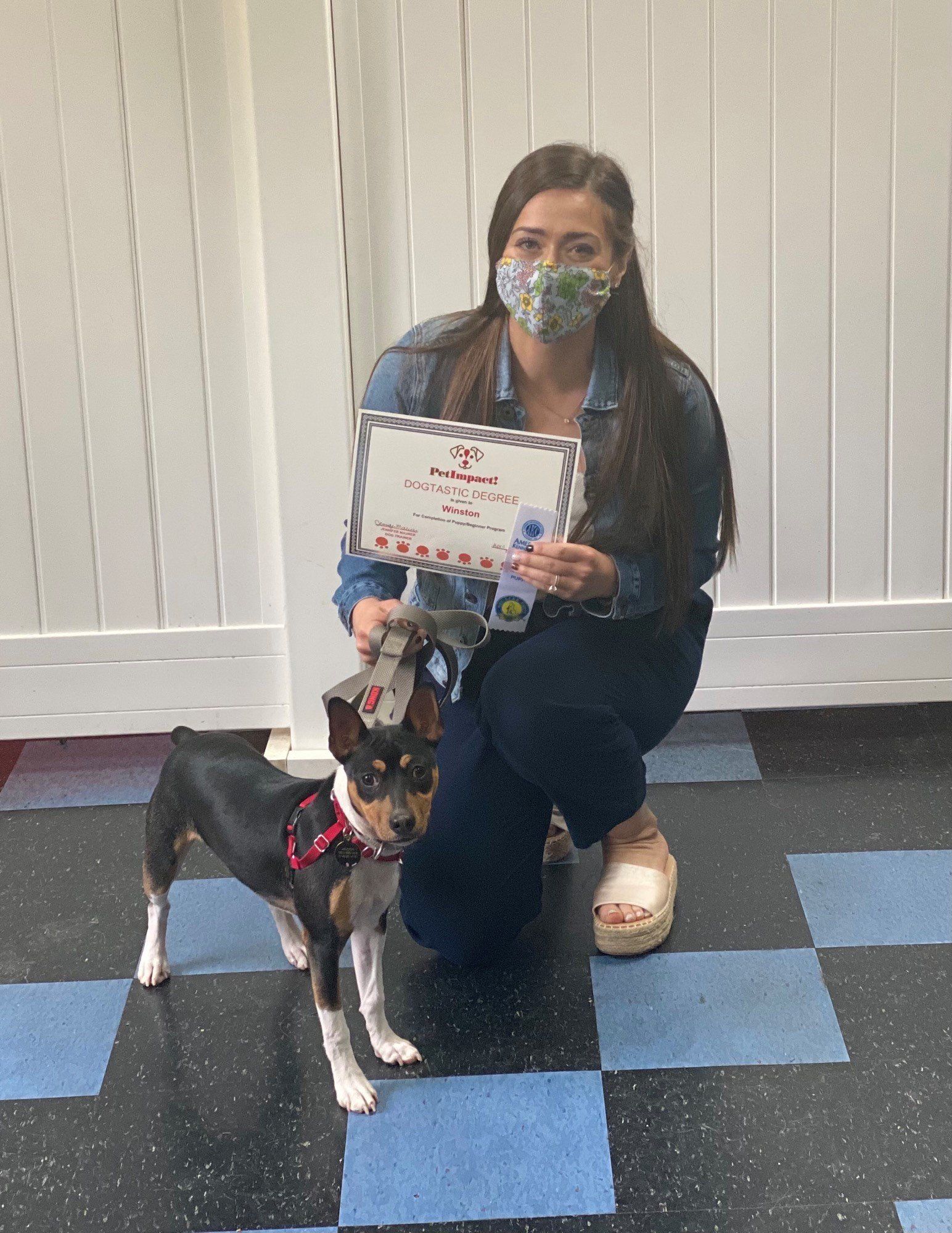 Woman with dog holds a certificate in front of a blue and black tiled floor, wearing a mask.