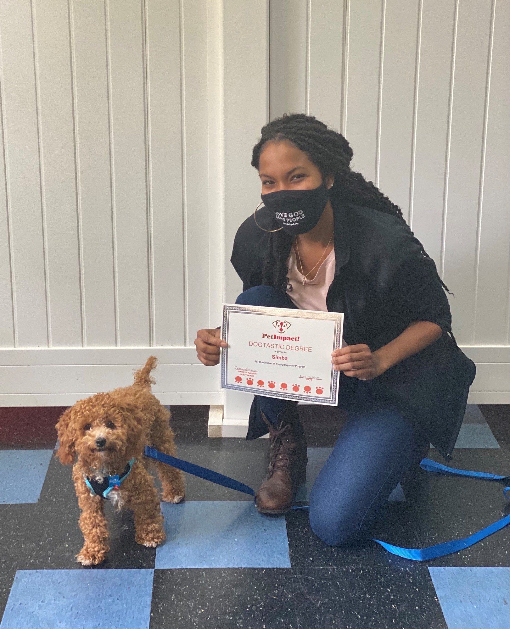 Woman kneels with dog. Woman holds certificate. Dog has leash and looks toward camera. Blue and black checkered floor.