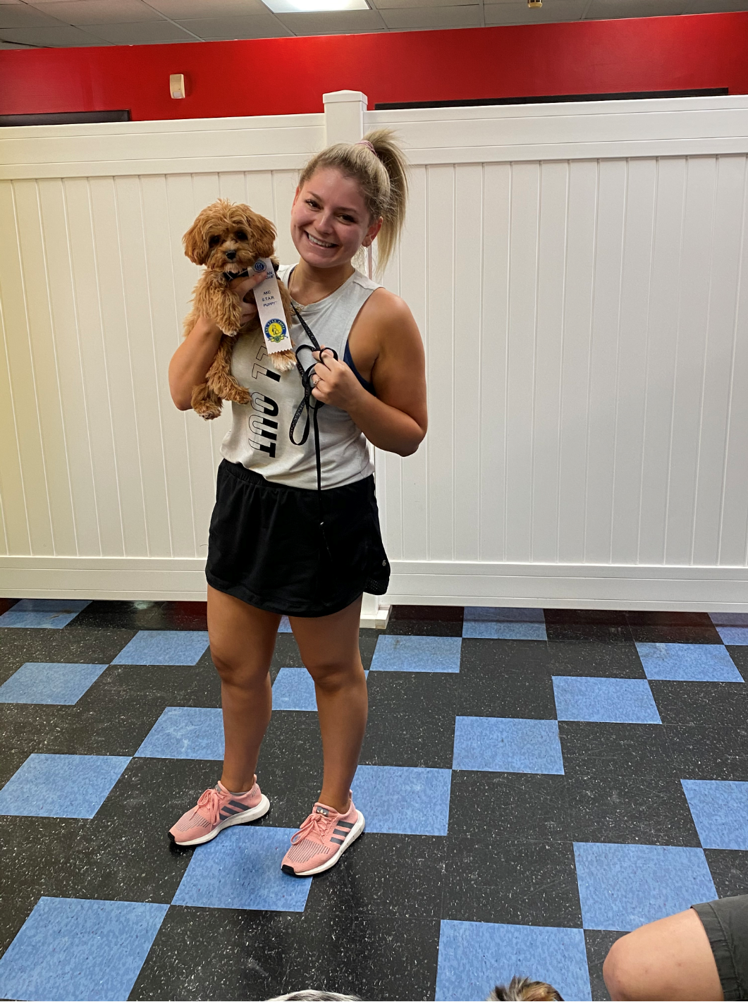 Woman holding a small brown dog, smiling. She is in a gym or indoor area with black and blue checkered floor.