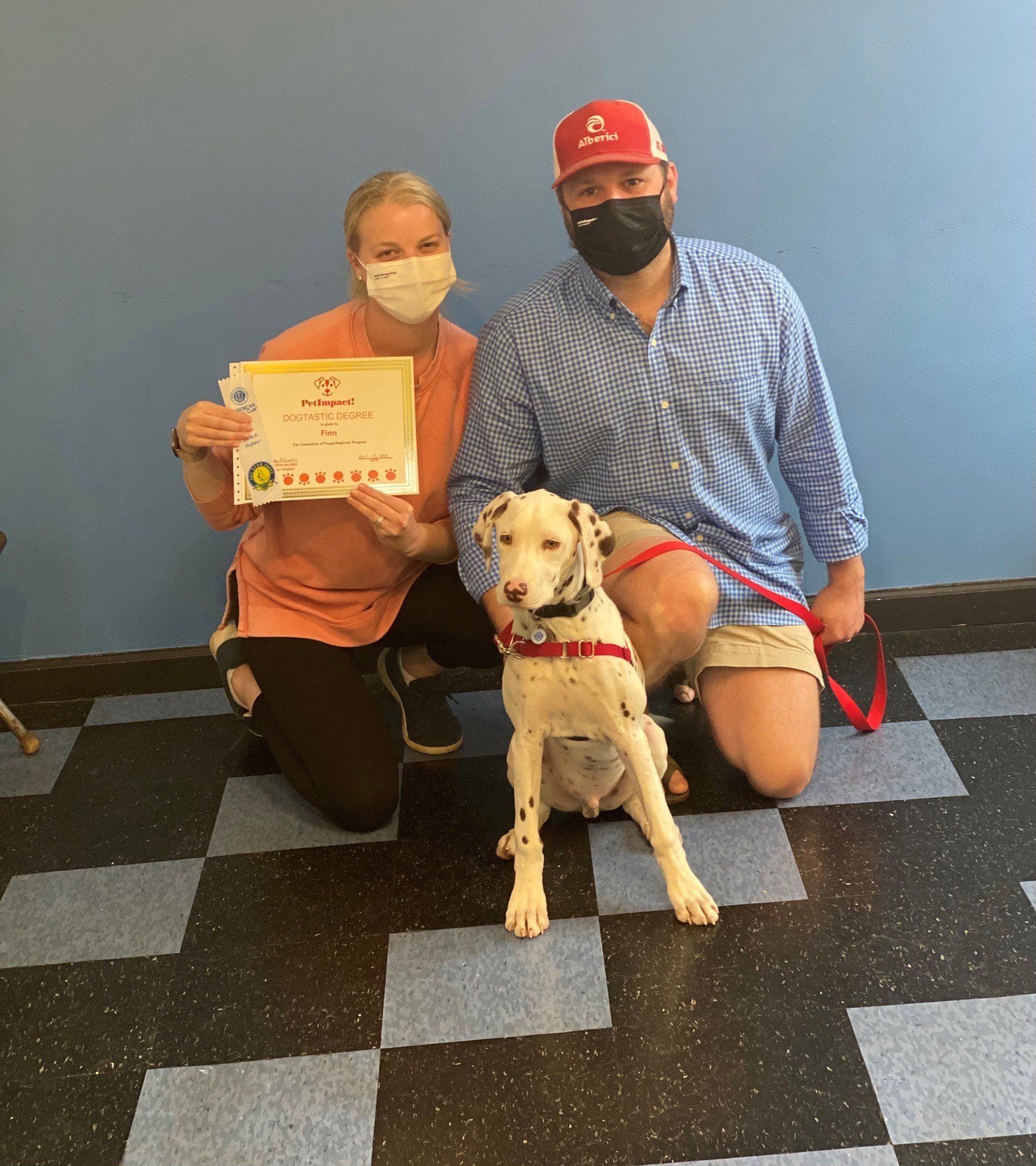 Dog with spots sits with two people, one holding certificate, all wearing masks in room with blue wall.