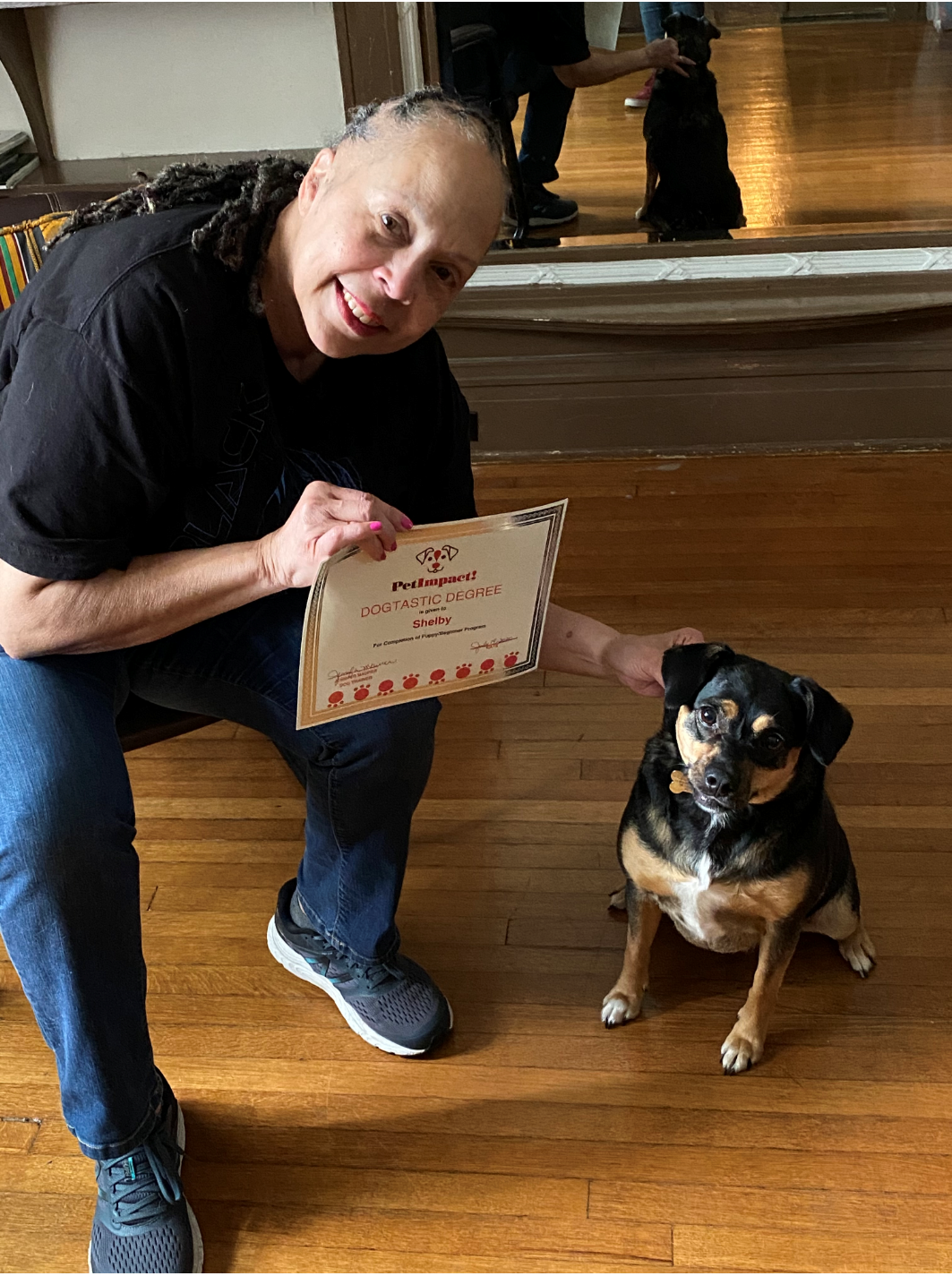 Person holding certificate, petting small dog; both smiling. Wooden floor, mirror reflection.