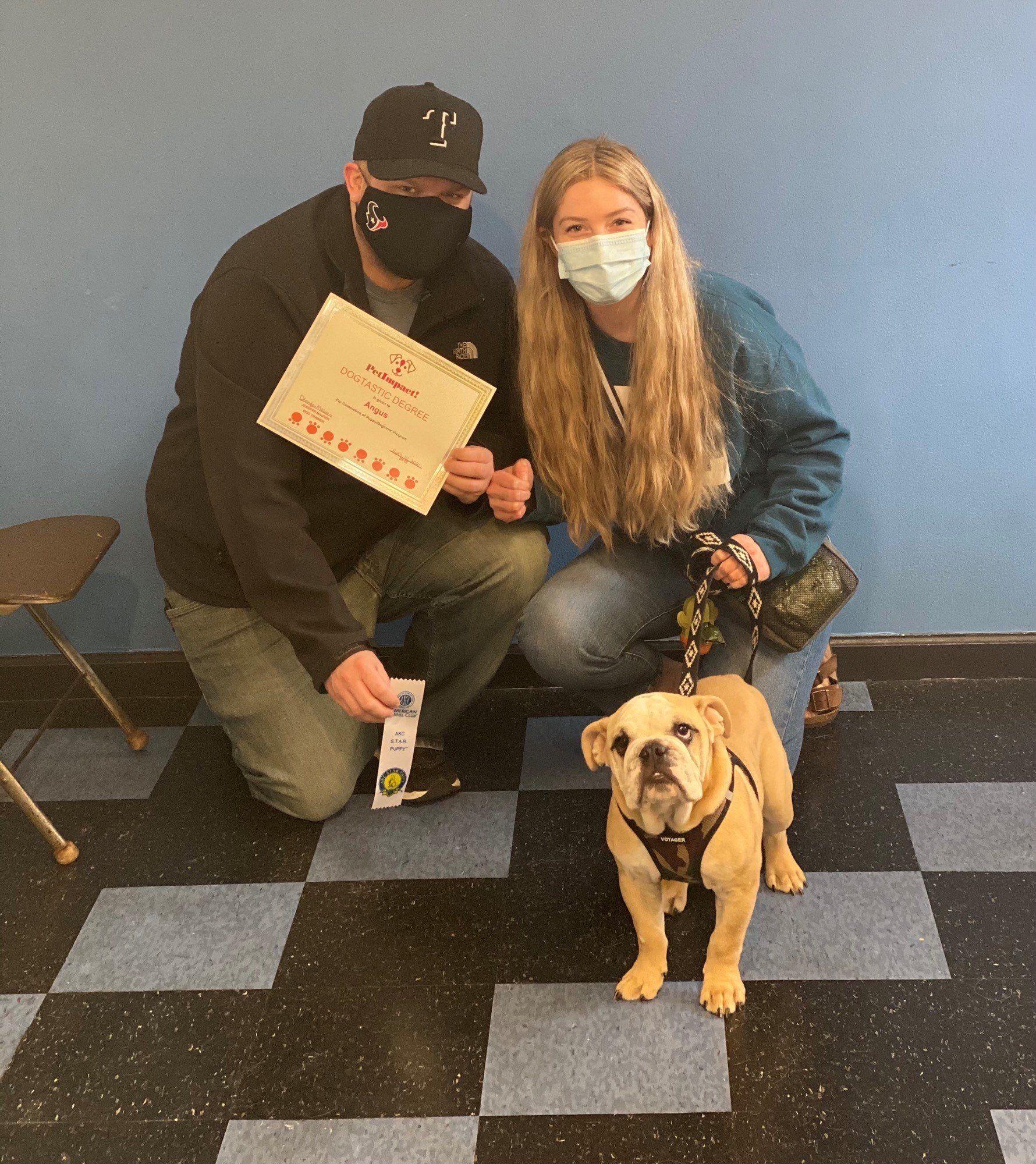 A couple kneels with their dog, holding a certificate. They are smiling and the dog is wearing a harness.