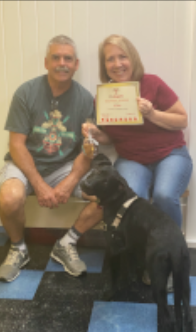 A couple sits with a black dog, holding a certificate. They smile. The floor is checkered blue and white.