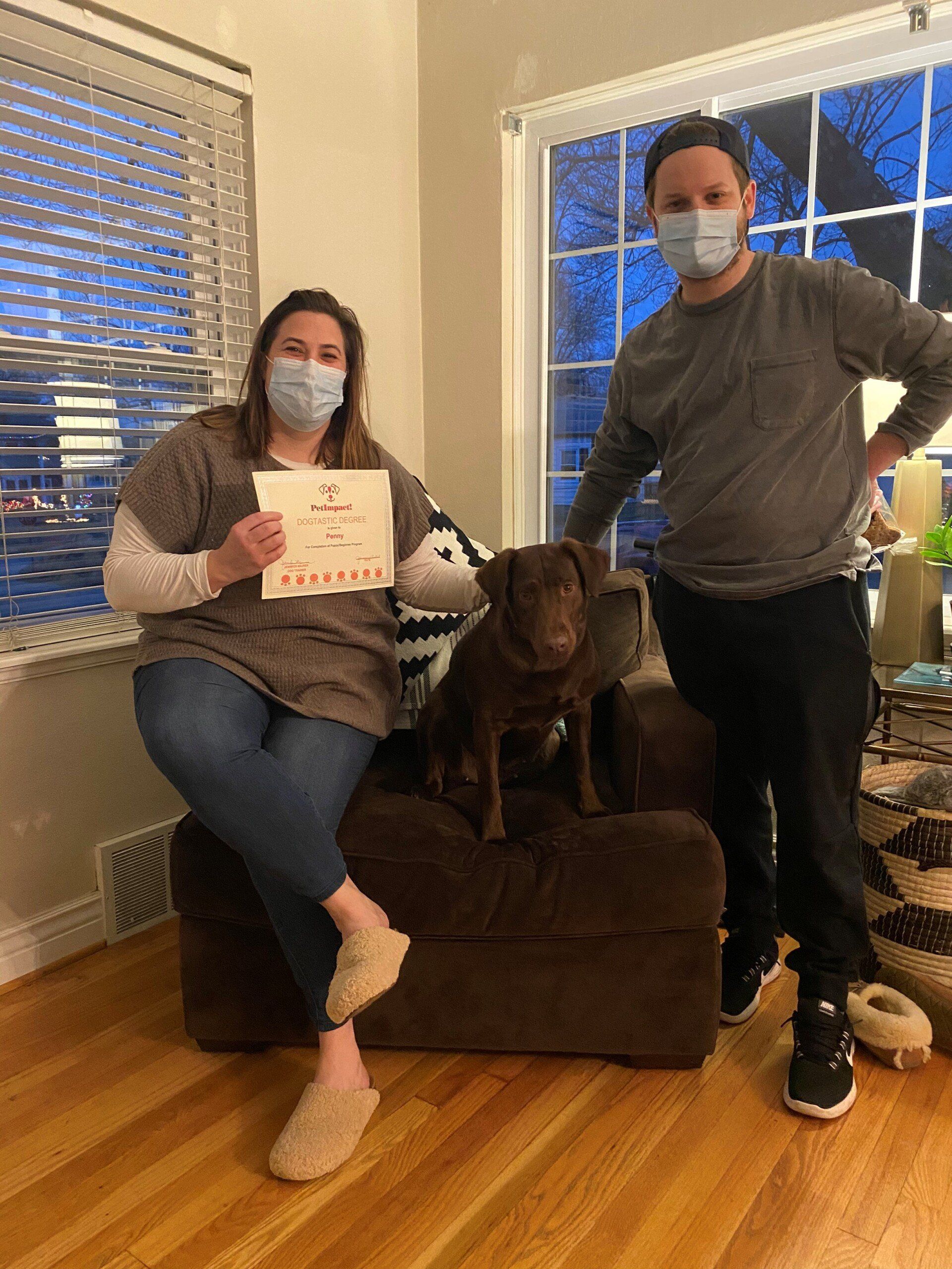 A couple and brown dog sit on a brown chair indoors. The woman holds a paper sign. All wear masks.