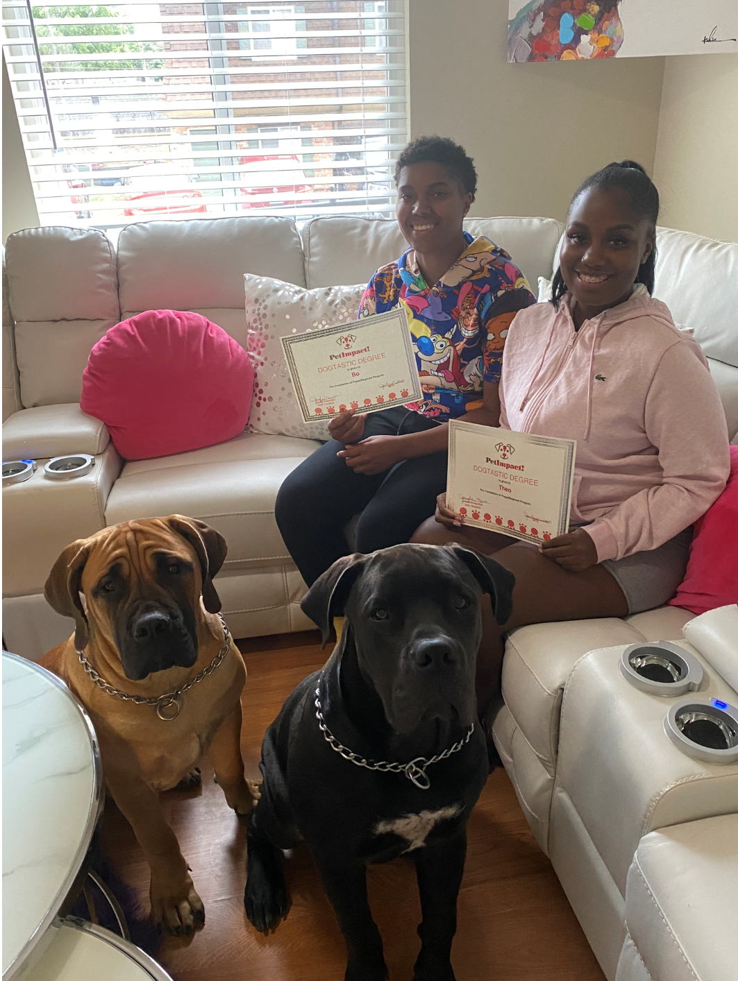 Two people on a couch with two dogs, holding signs. Pink and white decor, bright setting.