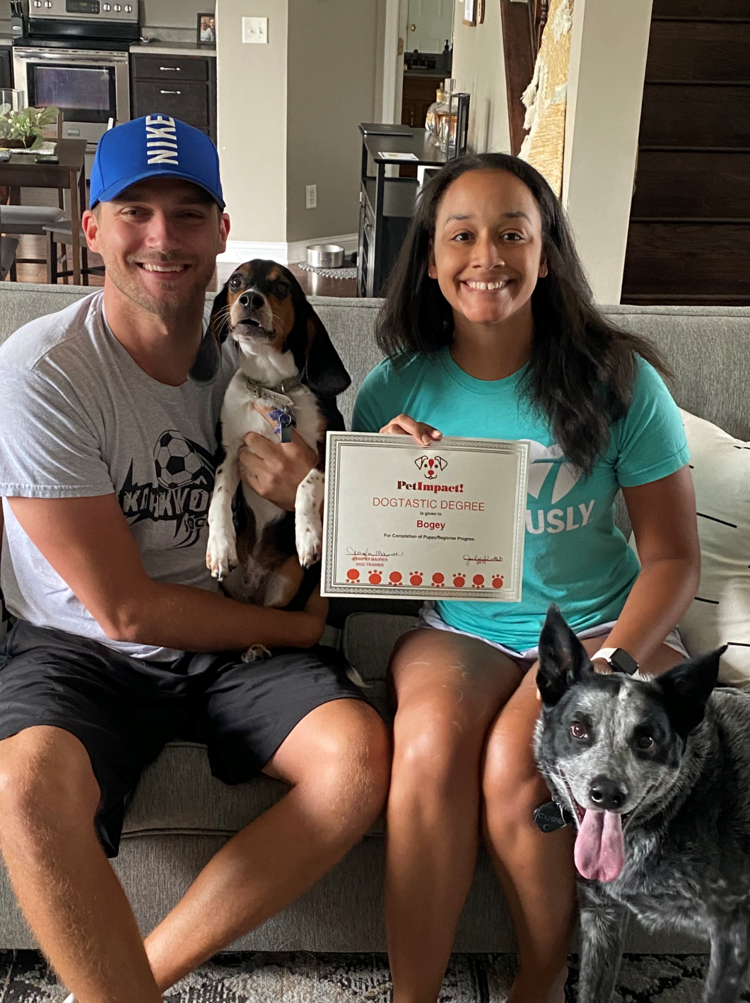 Couple on couch with two dogs and certificate; dog in man's arms, blue heeler on floor.