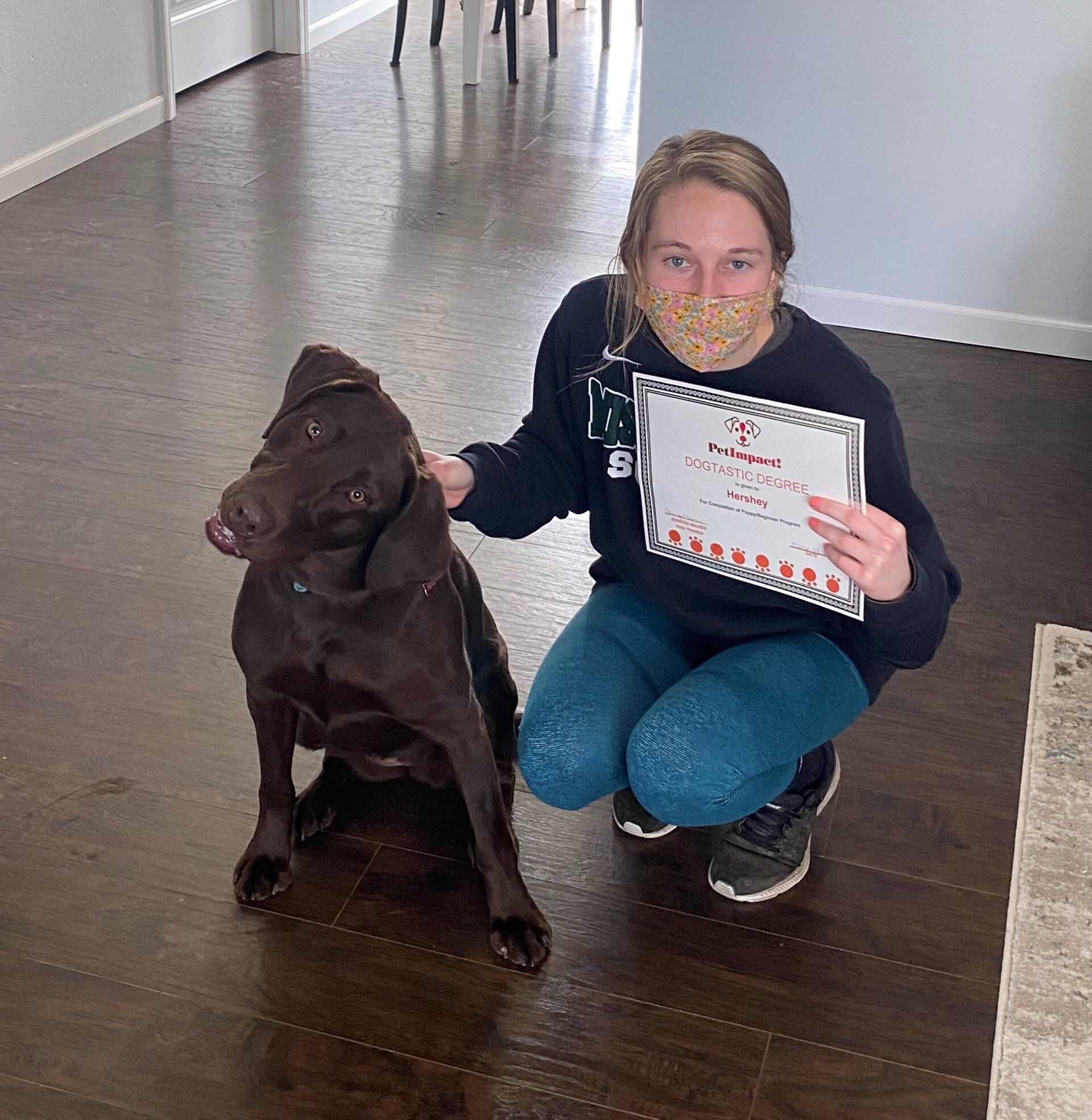Woman in mask, kneeling with brown dog; holding a certificate. Indoors, wooden floor.