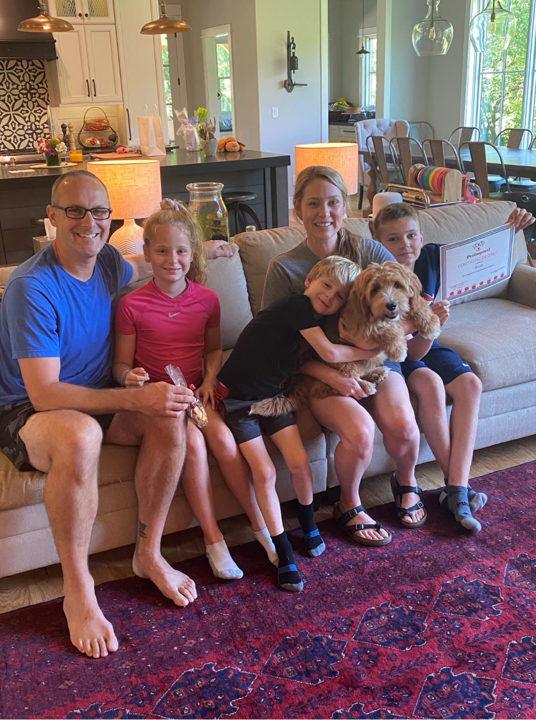 Family of five on a couch with a dog; sitting in a living room with sunlight.