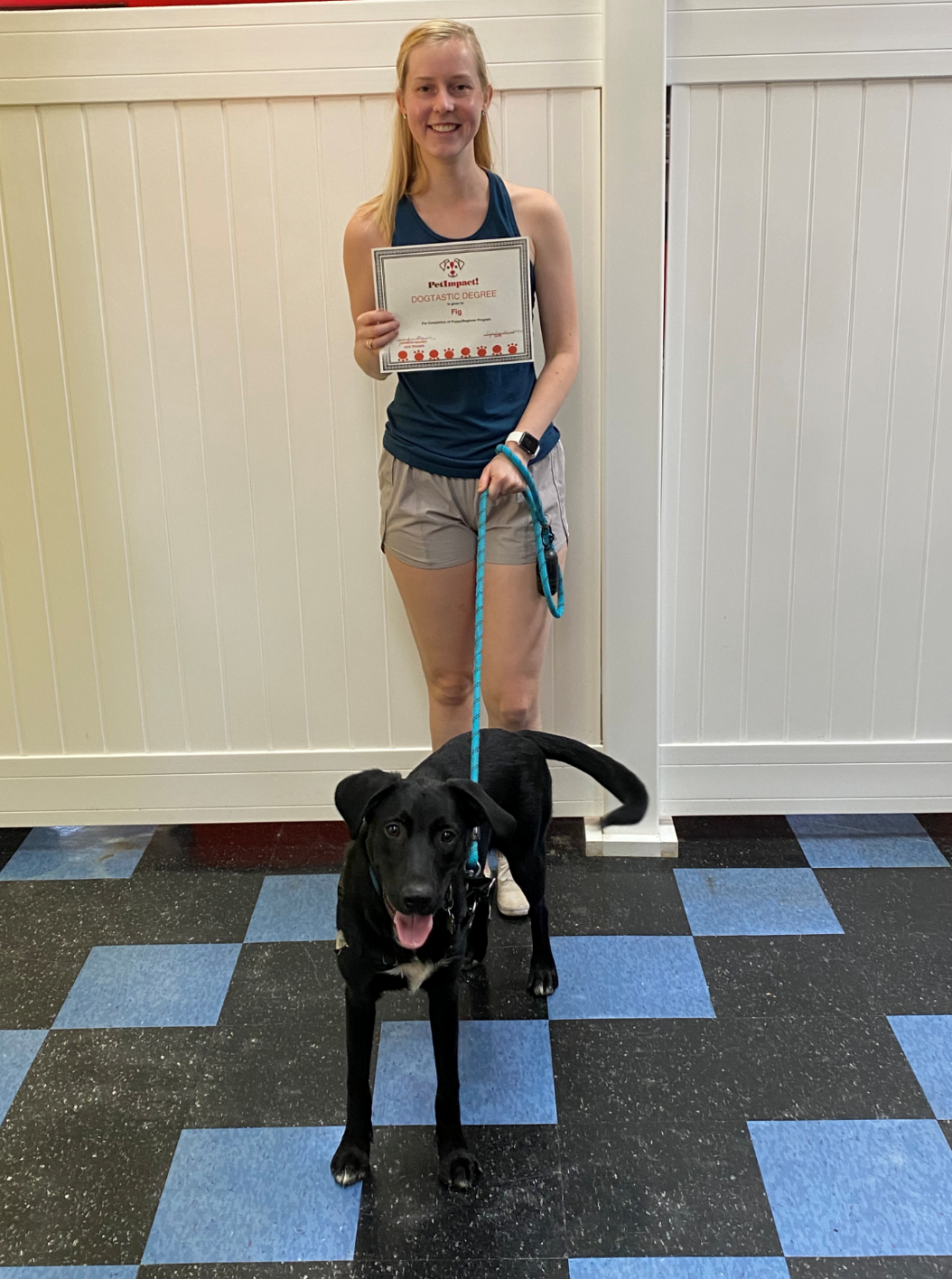 Woman and black dog in front of wall; woman holds certificate, dog on leash; they are smiling.
