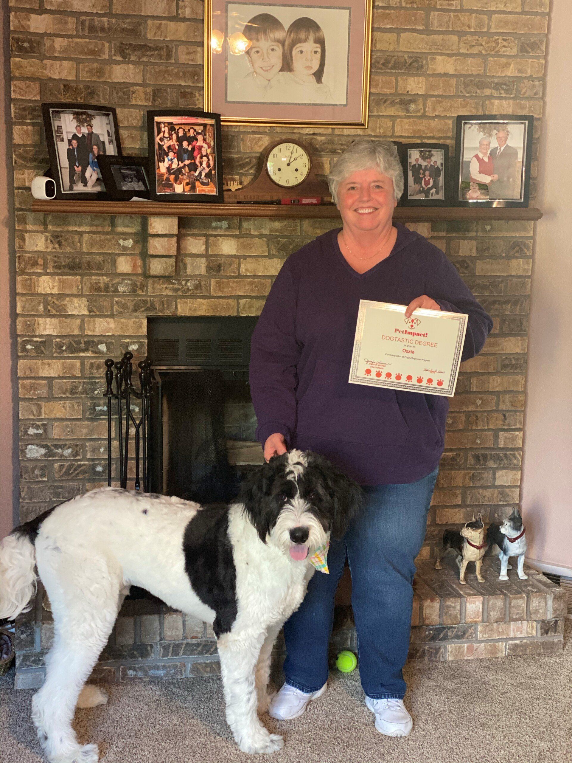Woman in purple shirt and jeans with a black and white dog, holding a sign near a fireplace.
