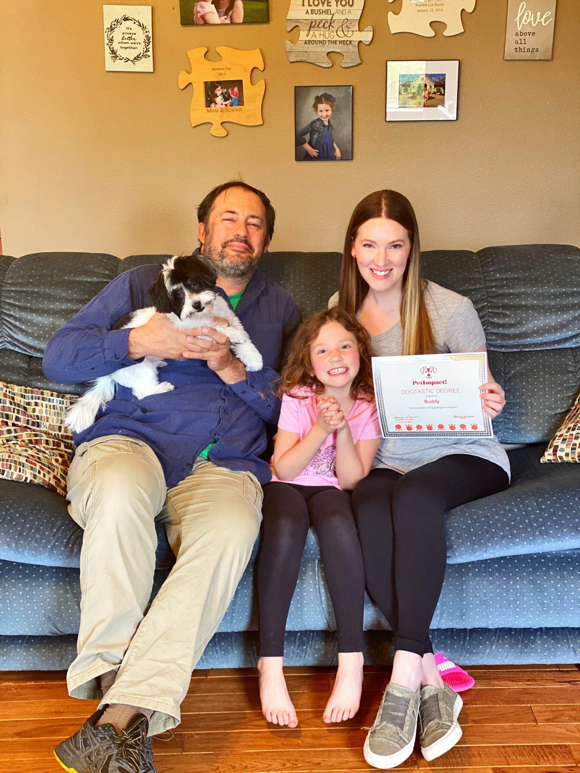 Family on a couch with a puppy, holding a certificate.  Smiling,  sitting in front of photos on a wall.