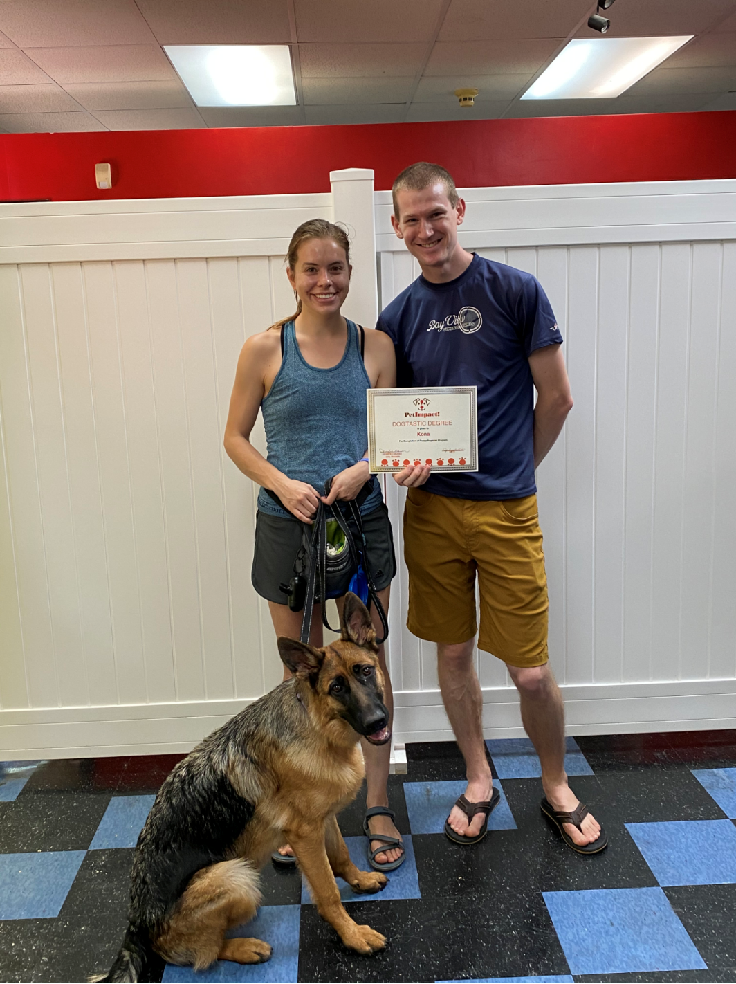 Woman, man, and German Shepherd pose with a certificate in a training facility. They stand in front of a white wall.