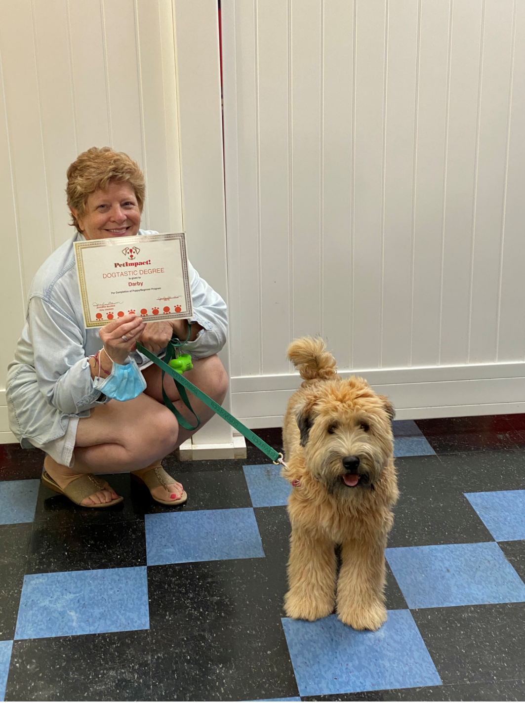 Woman in a striped shirt squats next to a fluffy tan dog, holding a certificate.  Both smile on a checkered floor.