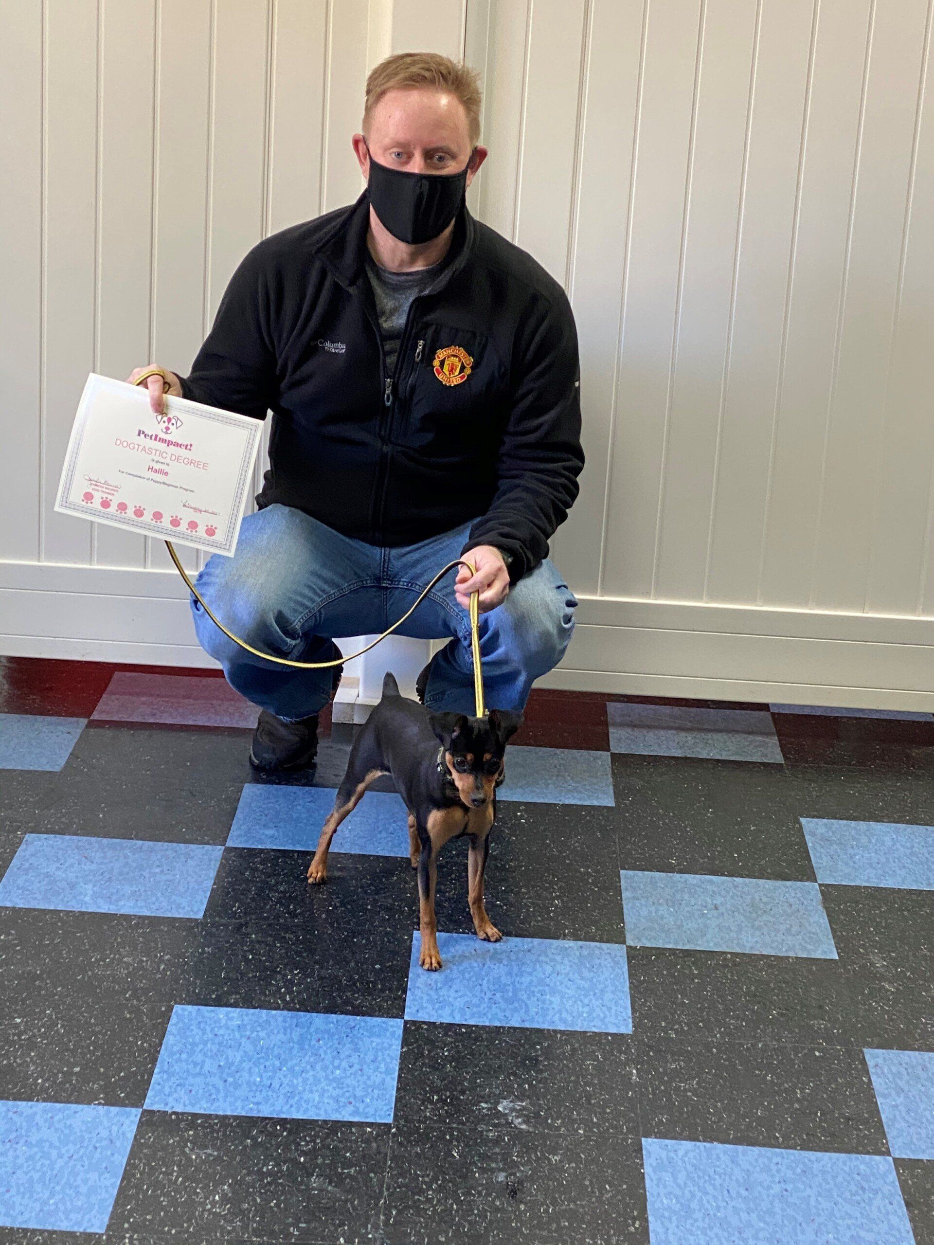 Man in black mask, crouched, holding a small dog on a leash and a paper; on blue and black tiled floor.