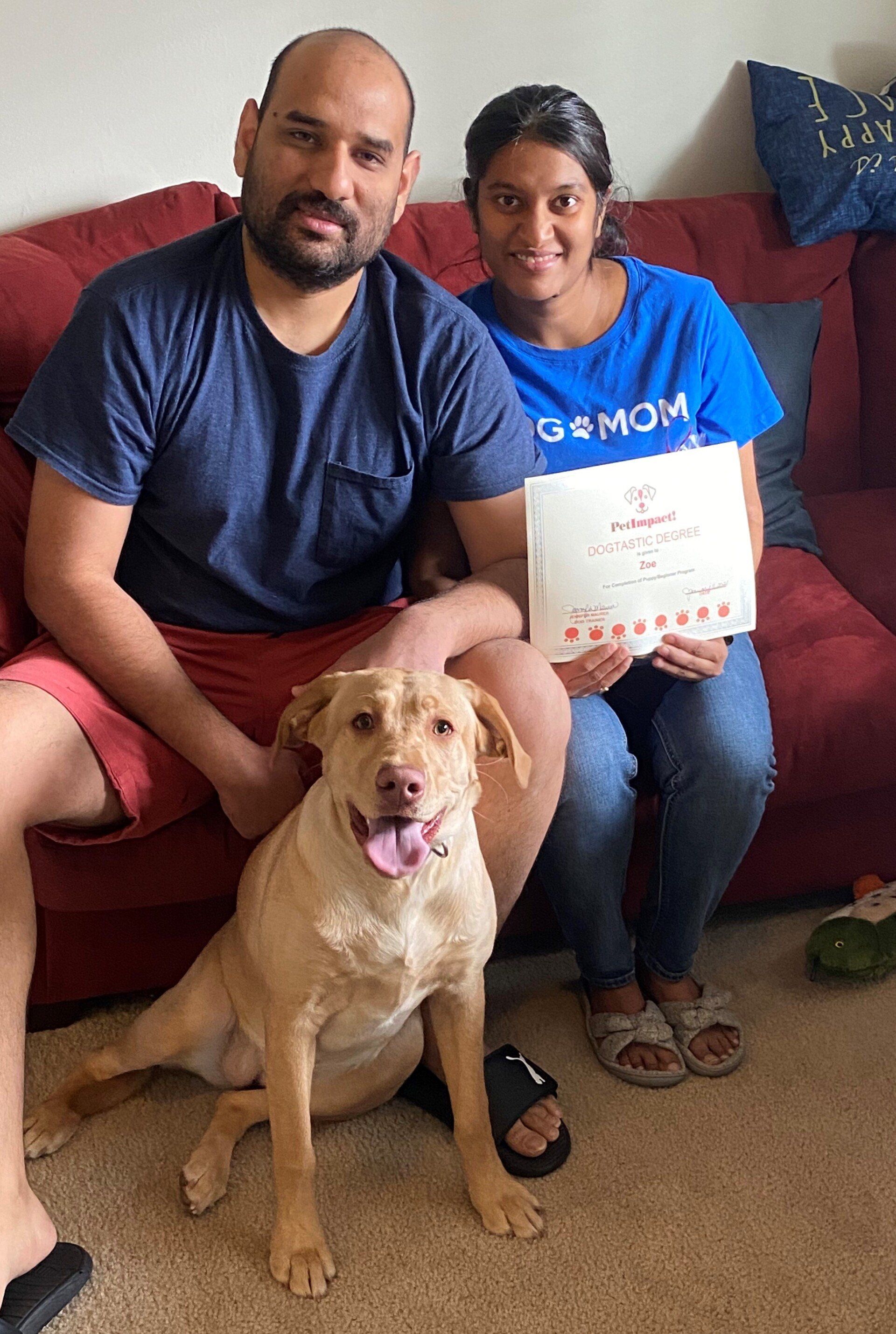 Couple and dog on a red couch. Woman holds a certificate. The dog is yellow with its tongue out.