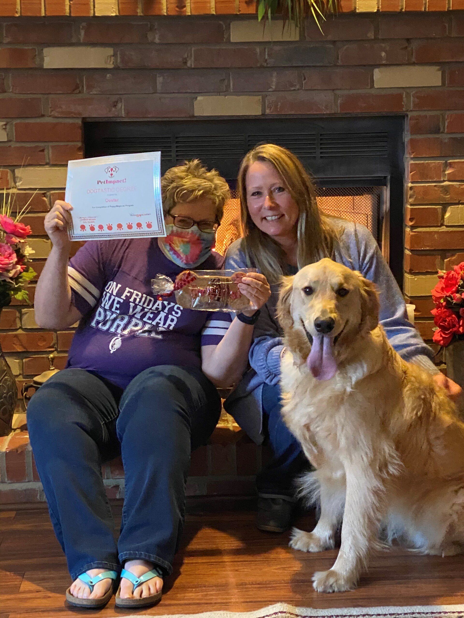 Two women and a golden retriever by a fireplace; one woman holds a certificate and container of treats.