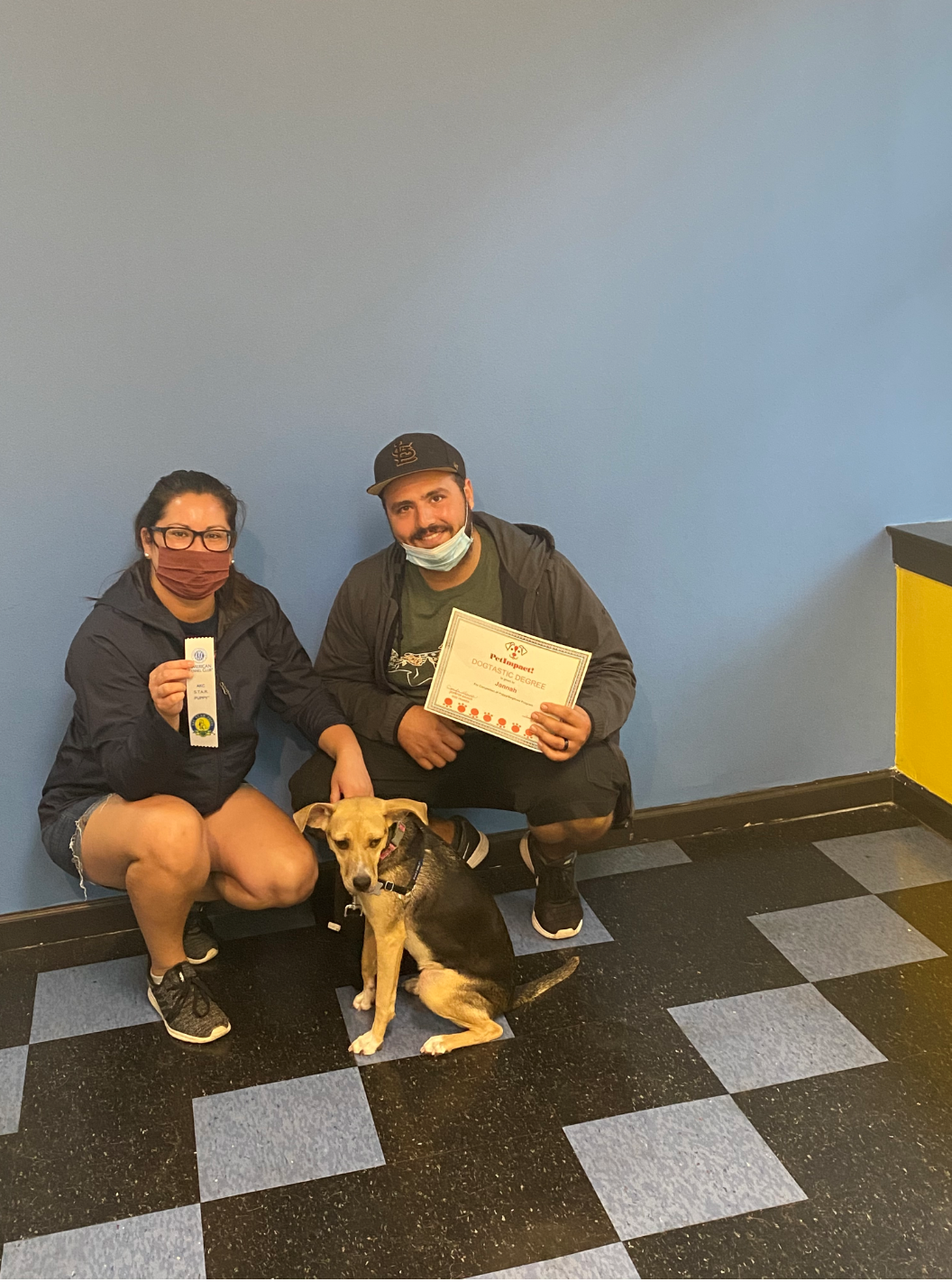 Two people and a dog crouch in front of a blue wall and checkered floor. One holds a certificate, the other a ribbon.