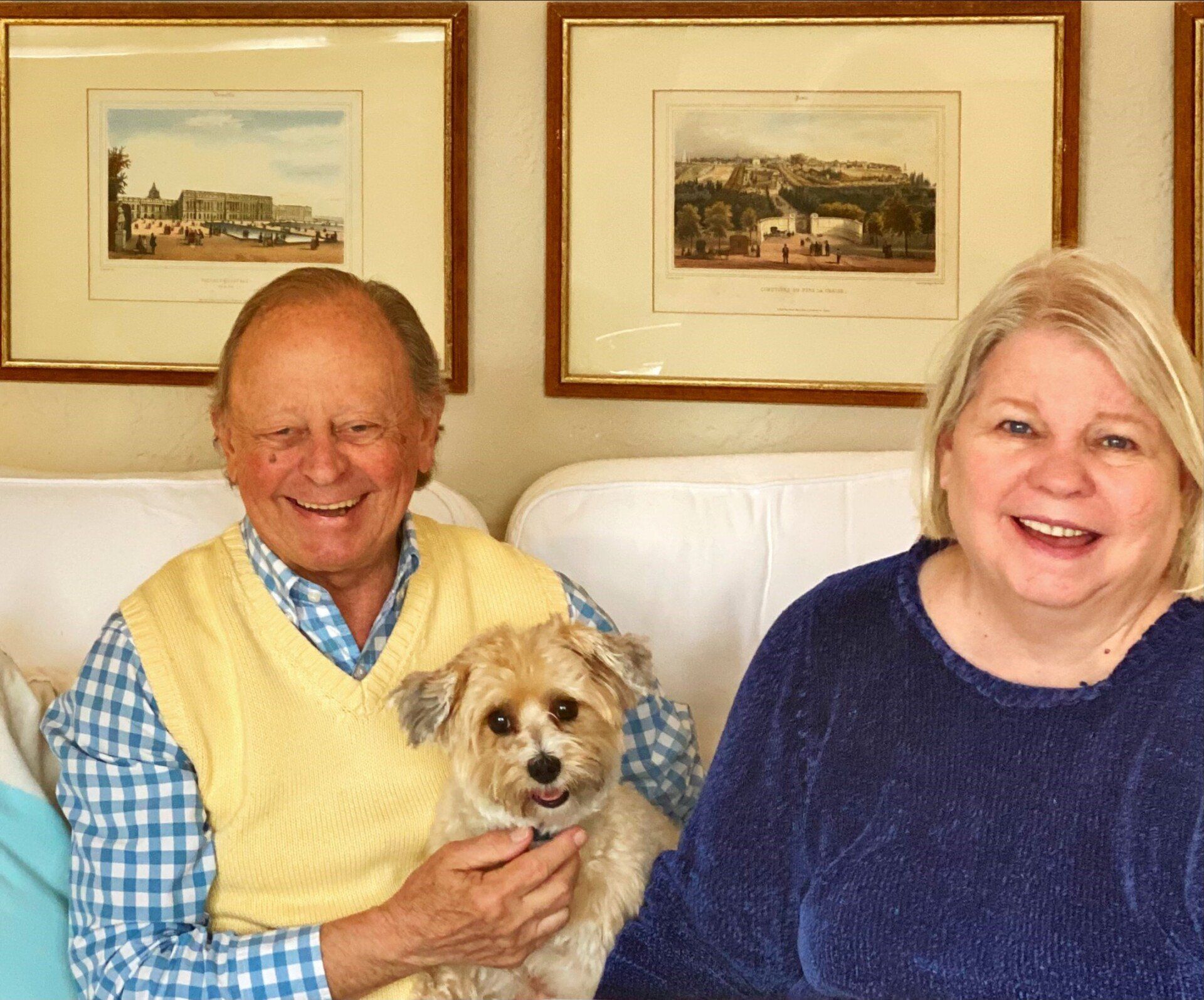 Smiling couple on a white couch with a small dog between them, framed artwork on the wall.